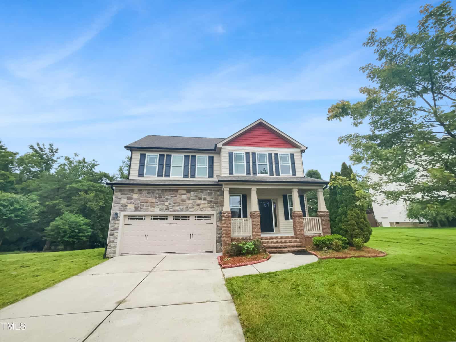 A two-story suburban house with stone and siding exterior, red gable, attached garage, front porch with columns, and a neatly manicured lawn under a clear blue sky.