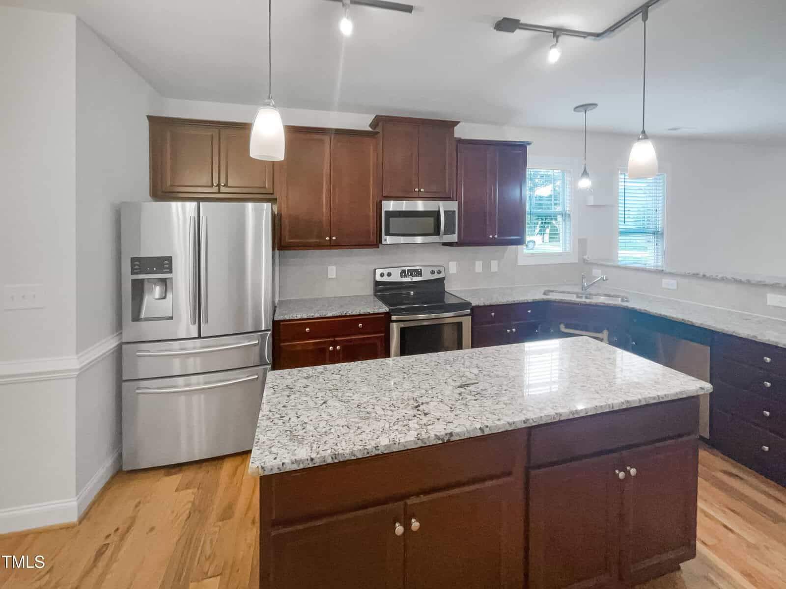 Modern kitchen with stainless steel appliances, dark wood cabinets, granite countertops, an island with storage, pendant lights, hardwood floors, and windows letting in natural light.