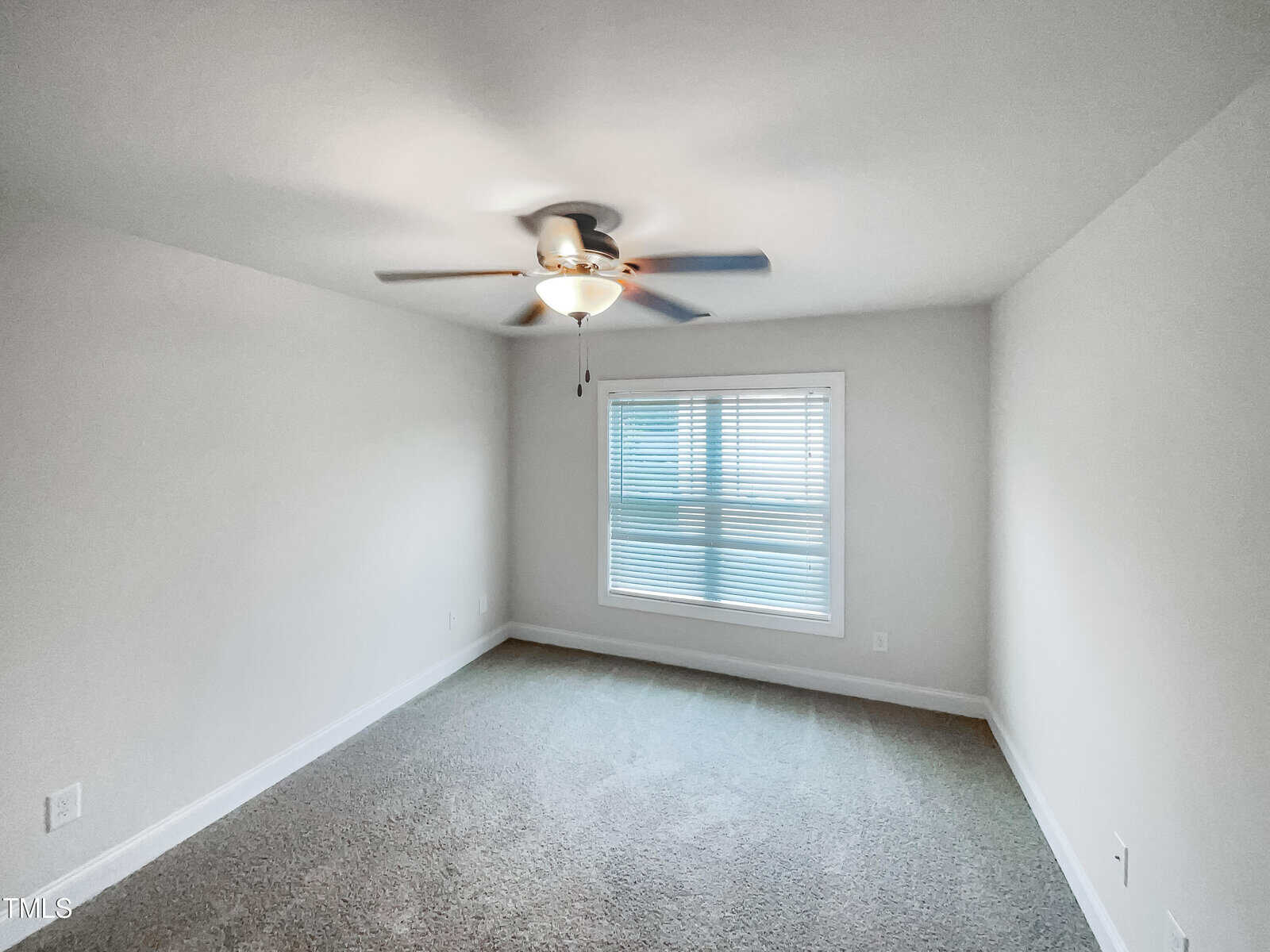 A bright, empty room with light gray carpet, white walls, a window with blinds, and a ceiling fan with a light fixture in the center of the ceiling.