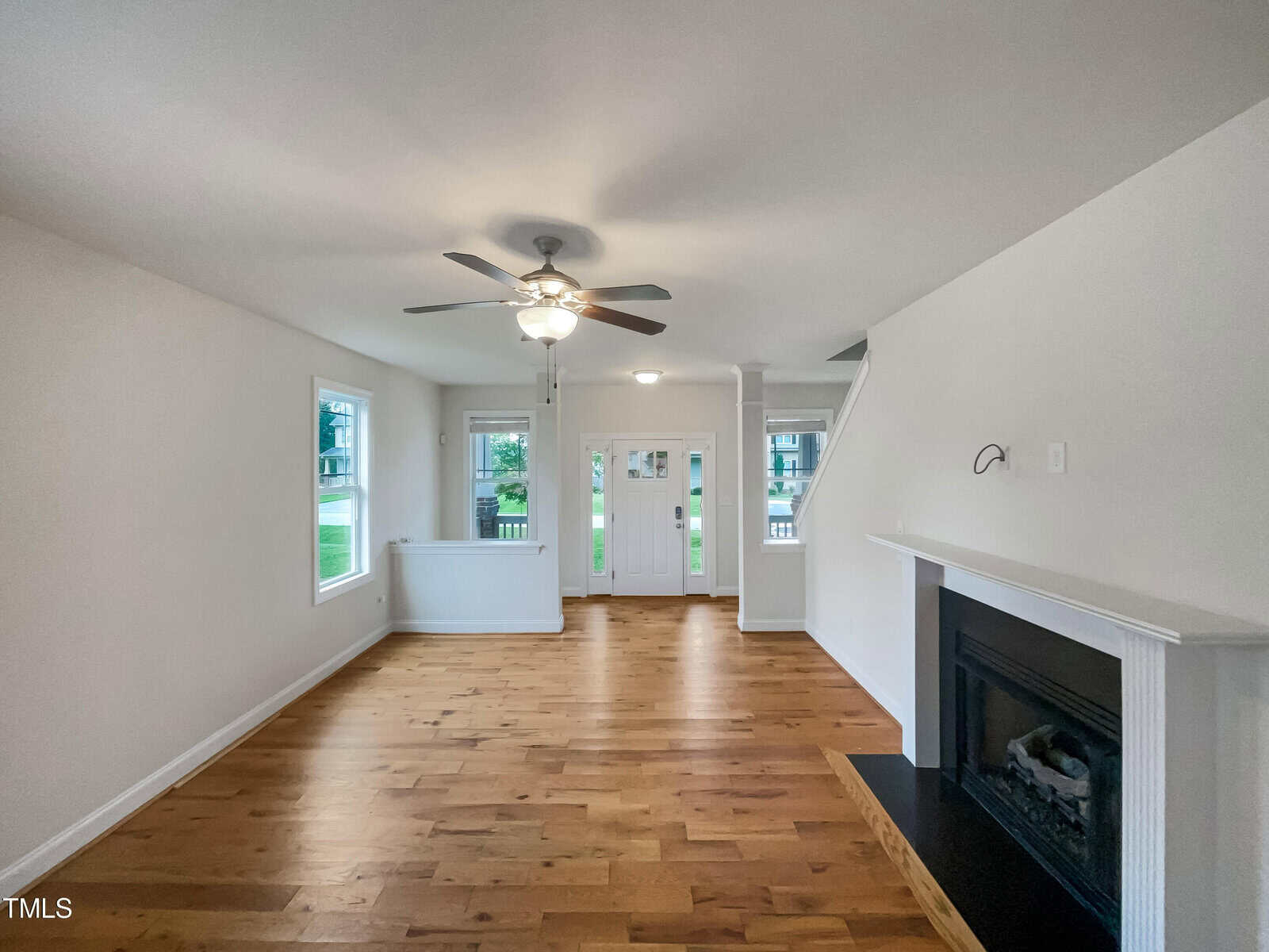 A bright, empty living room with light walls, hardwood floors, a ceiling fan, a fireplace on the right, and large windows near a white front door with sidelights. A staircase is visible to the right of the entrance.