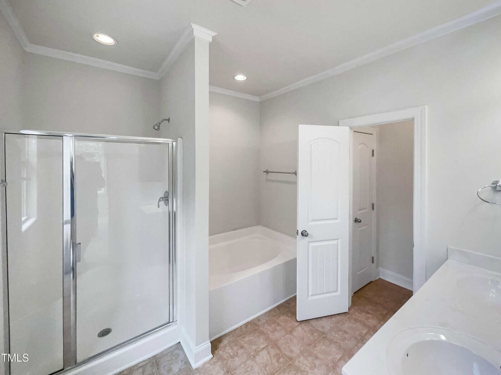 Bright bathroom with a glass shower, white bathtub in the corner, open white door, double sink vanity with mirror, beige tiled floor, and light-colored walls.