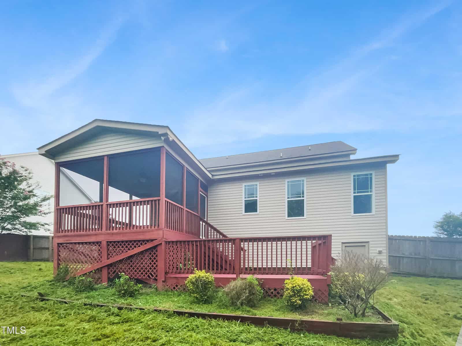 A beige house with a spacious backyard features a red screened-in porch and deck, surrounded by green grass, small bushes, and a wooden fence, under a blue sky.