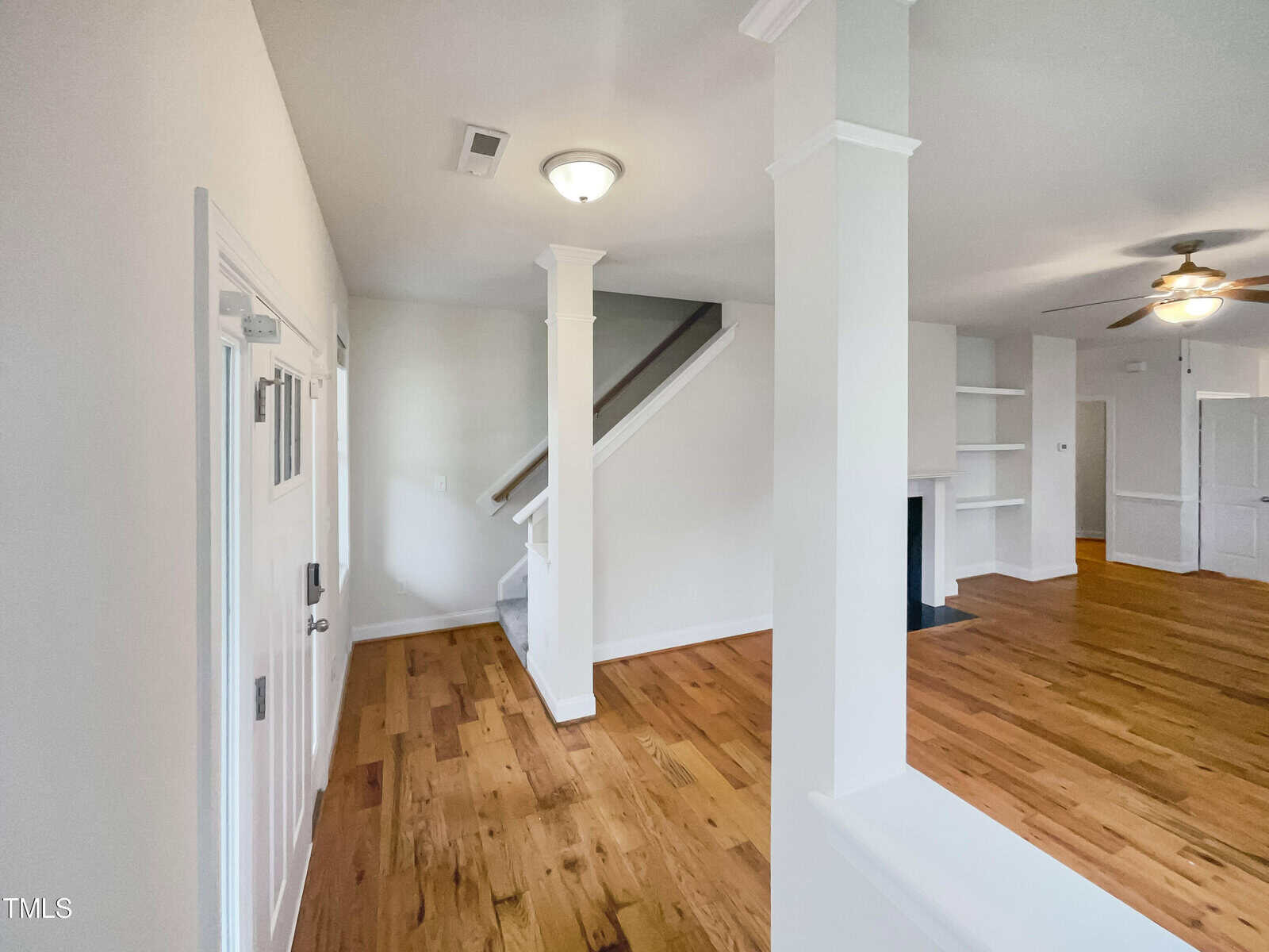 A bright, open interior with hardwood floors, white walls, a staircase with a white railing, built-in shelves, and a ceiling fan in the living area. Natural light fills the space.