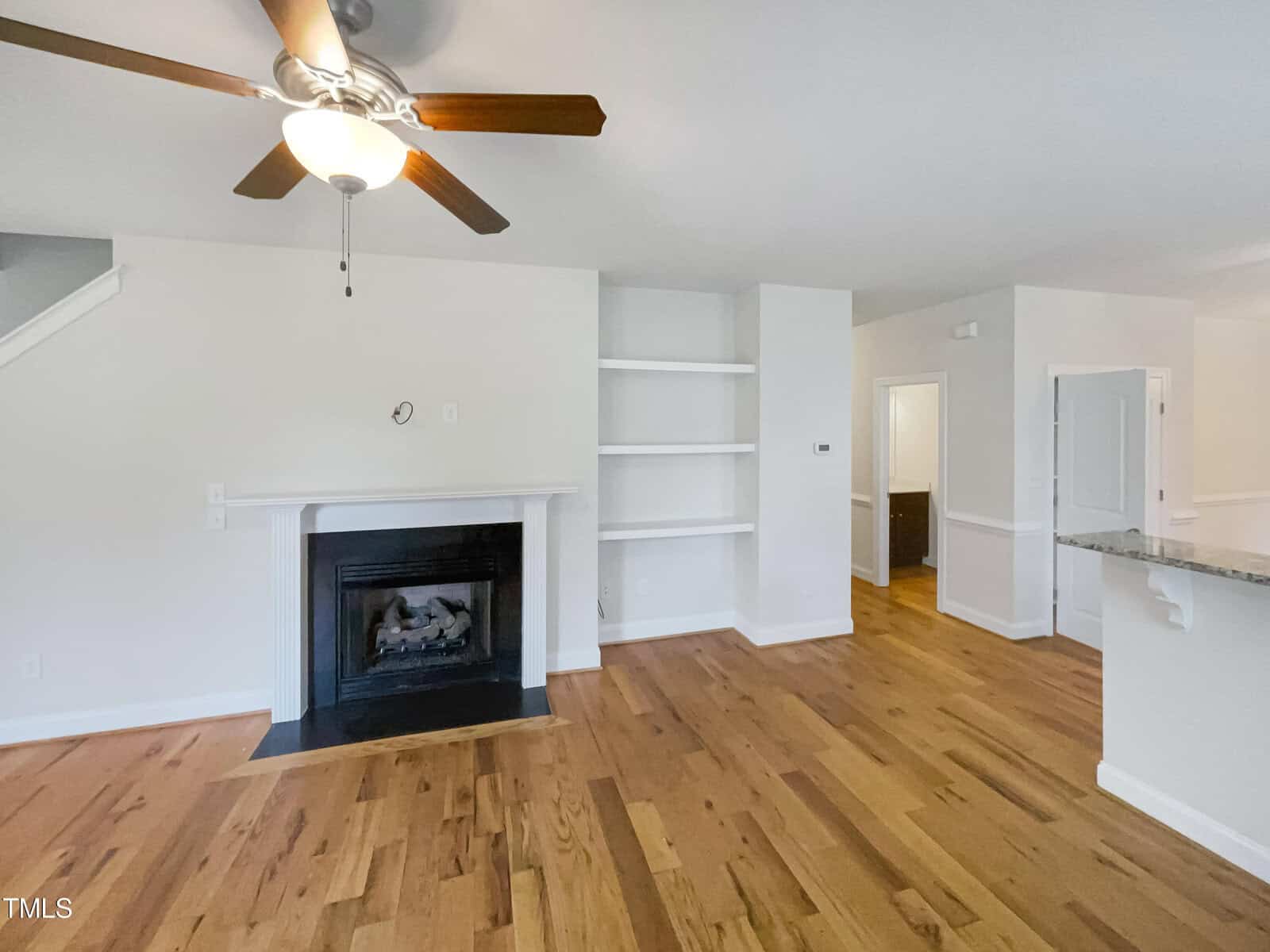 A bright living room with hardwood floors, a ceiling fan, a fireplace with mantel, built-in shelves, and white walls. An open doorway leads to another room, and a partial kitchen counter is visible.