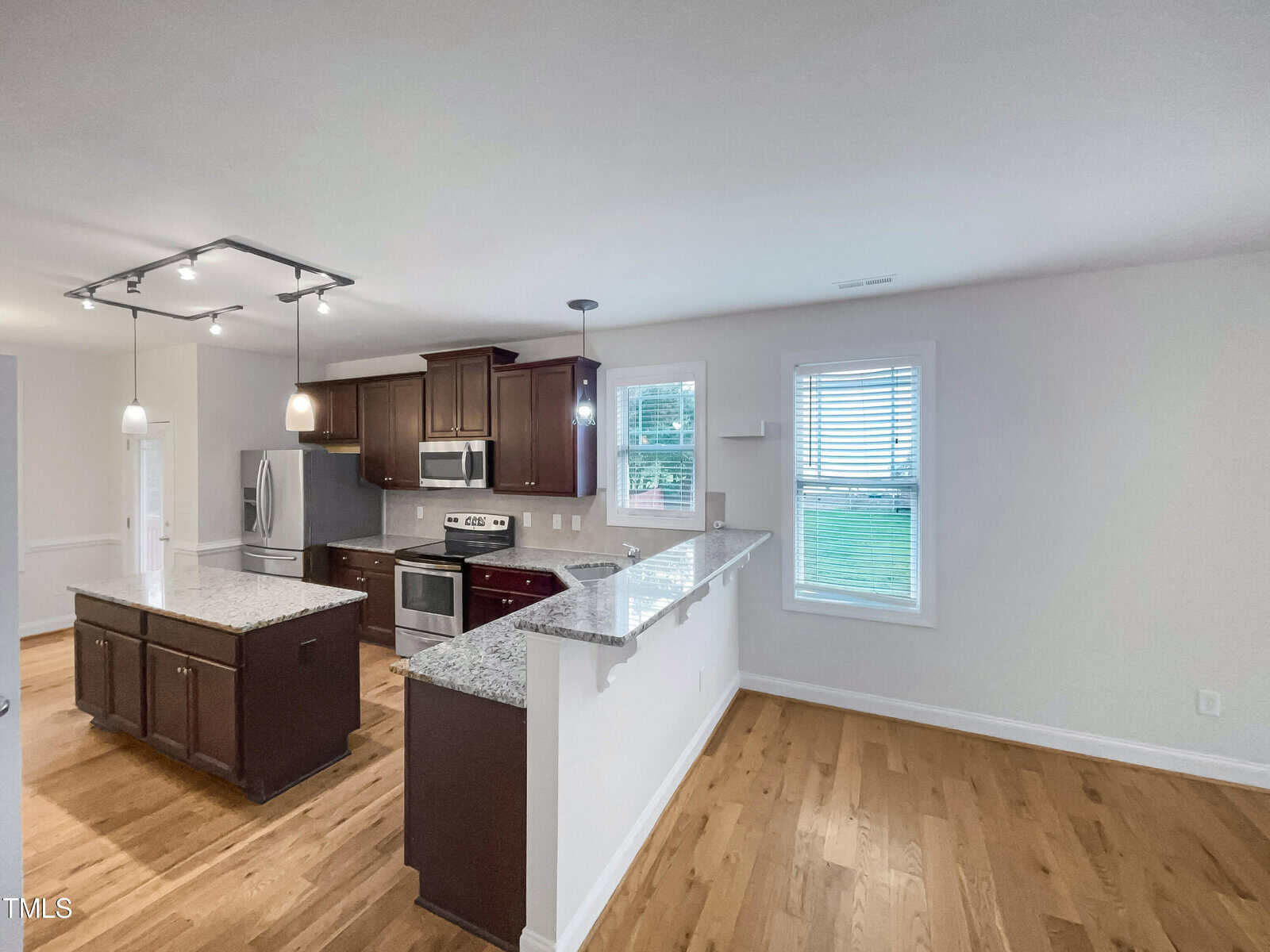 Modern kitchen with dark wood cabinets, stainless steel appliances, granite countertops, a center island, and light hardwood floors. Two windows and pendant lights provide natural and artificial light.