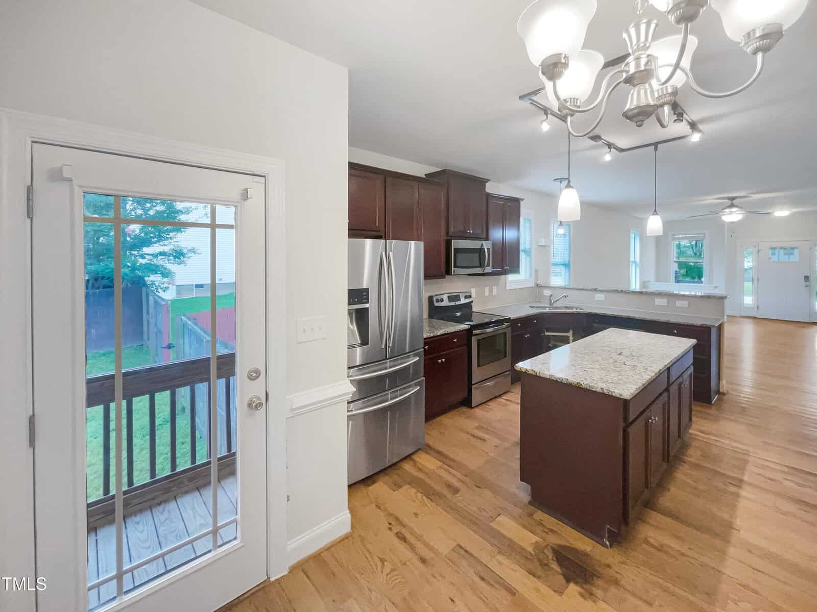 Modern kitchen with stainless steel appliances, dark wood cabinets, a central island with granite countertop, and hardwood floors. A glass door leads to a wooden deck and backyard. Open layout connects to living area.