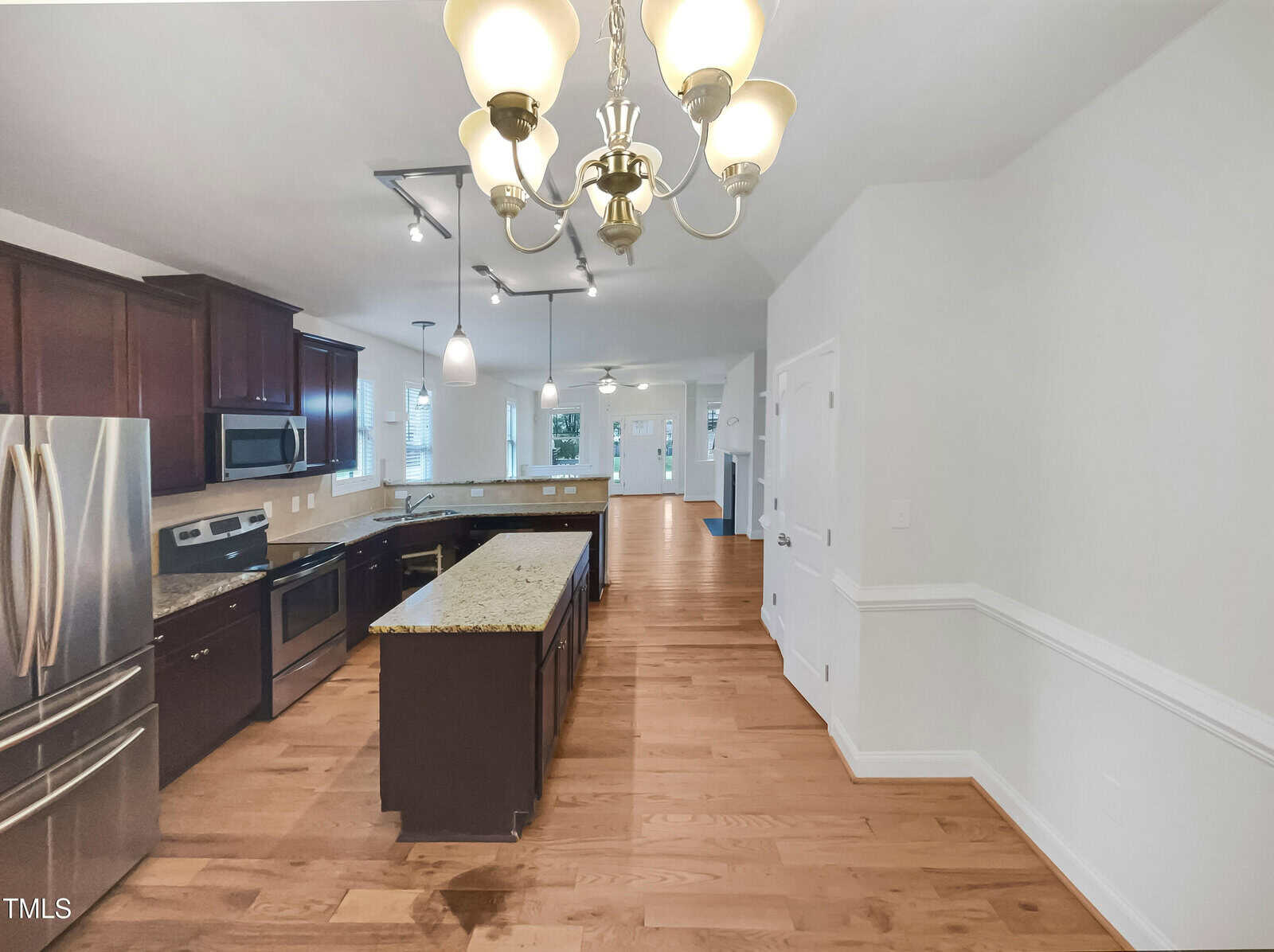 Spacious kitchen with dark wood cabinets, stainless steel appliances, granite countertops, island, light fixtures, hardwood floors, and open layout leading to a bright living area.
