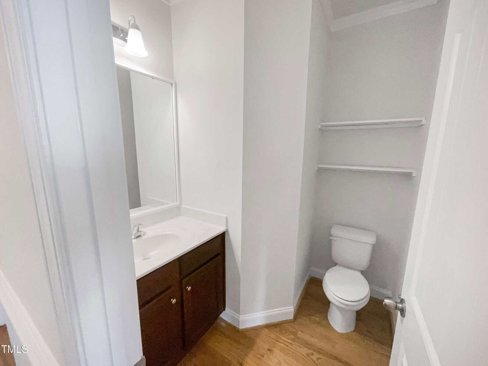 A small bathroom with a white toilet, a sink with a dark wood cabinet, a large mirror, white walls, hardwood floor, and three white shelves above the toilet.