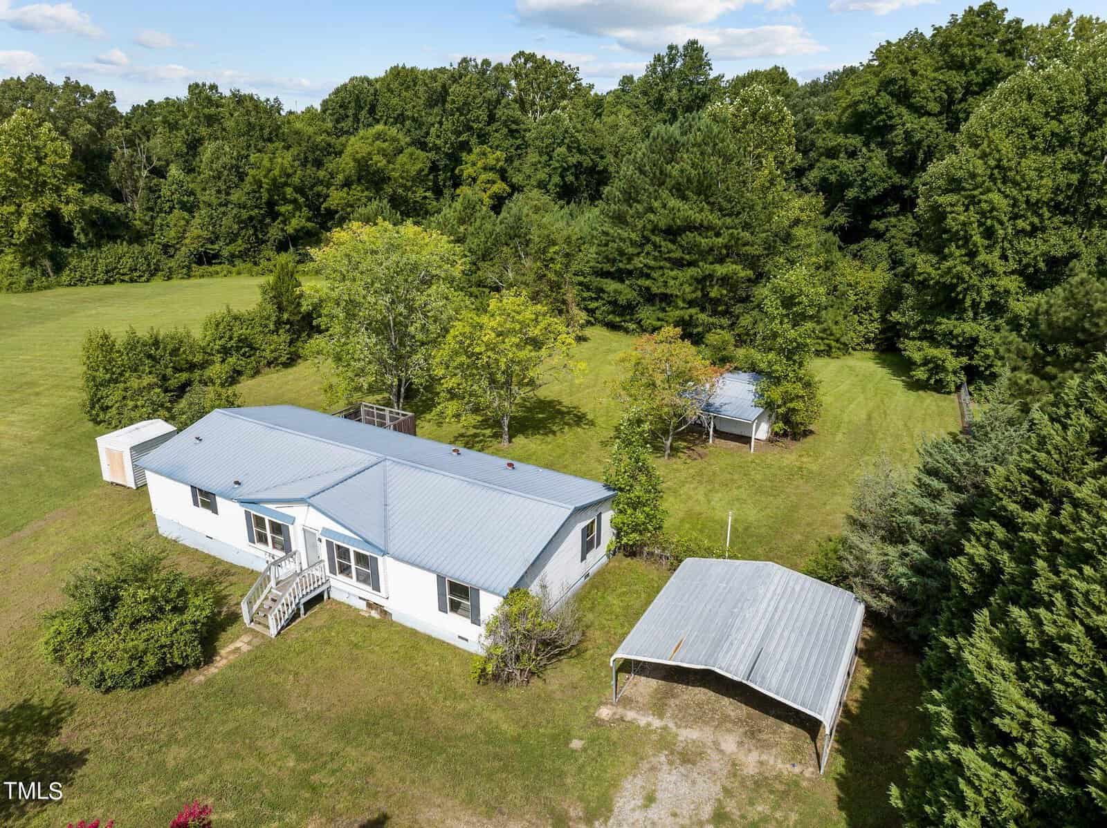 An aerial view reveals a white mobile home with a blue roof, nestled in a spacious grassy area along 4651 NC Highway 96 near Oxford. Surrounded by trees, the scene features a metal carport to the right and a small white shed visible in the distance.