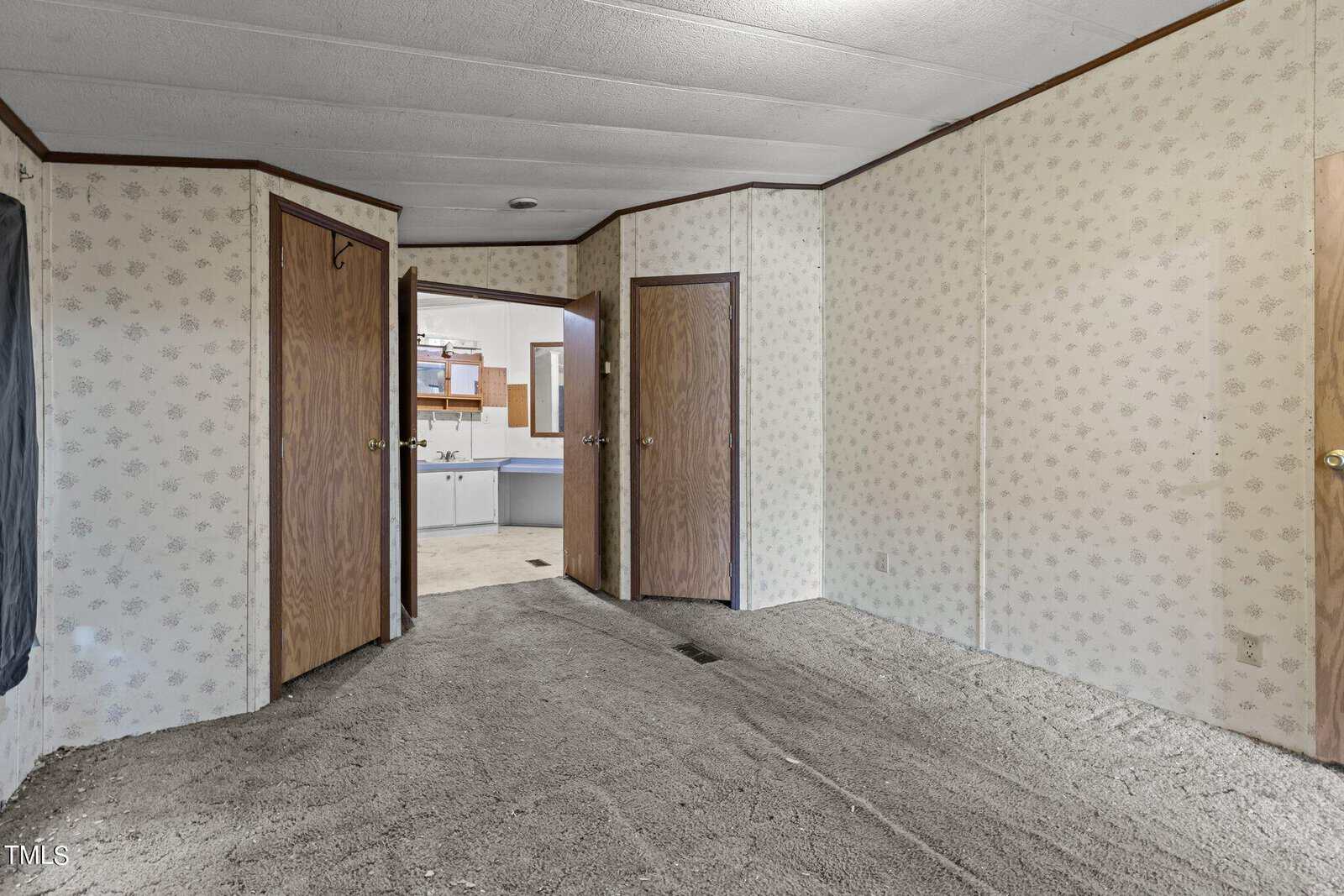 An empty room with patterned wallpaper and carpet flooring. The room has two wooden doors and a door leading to a kitchen area. The kitchen has visible cabinets and a sink. The room is well-lit.