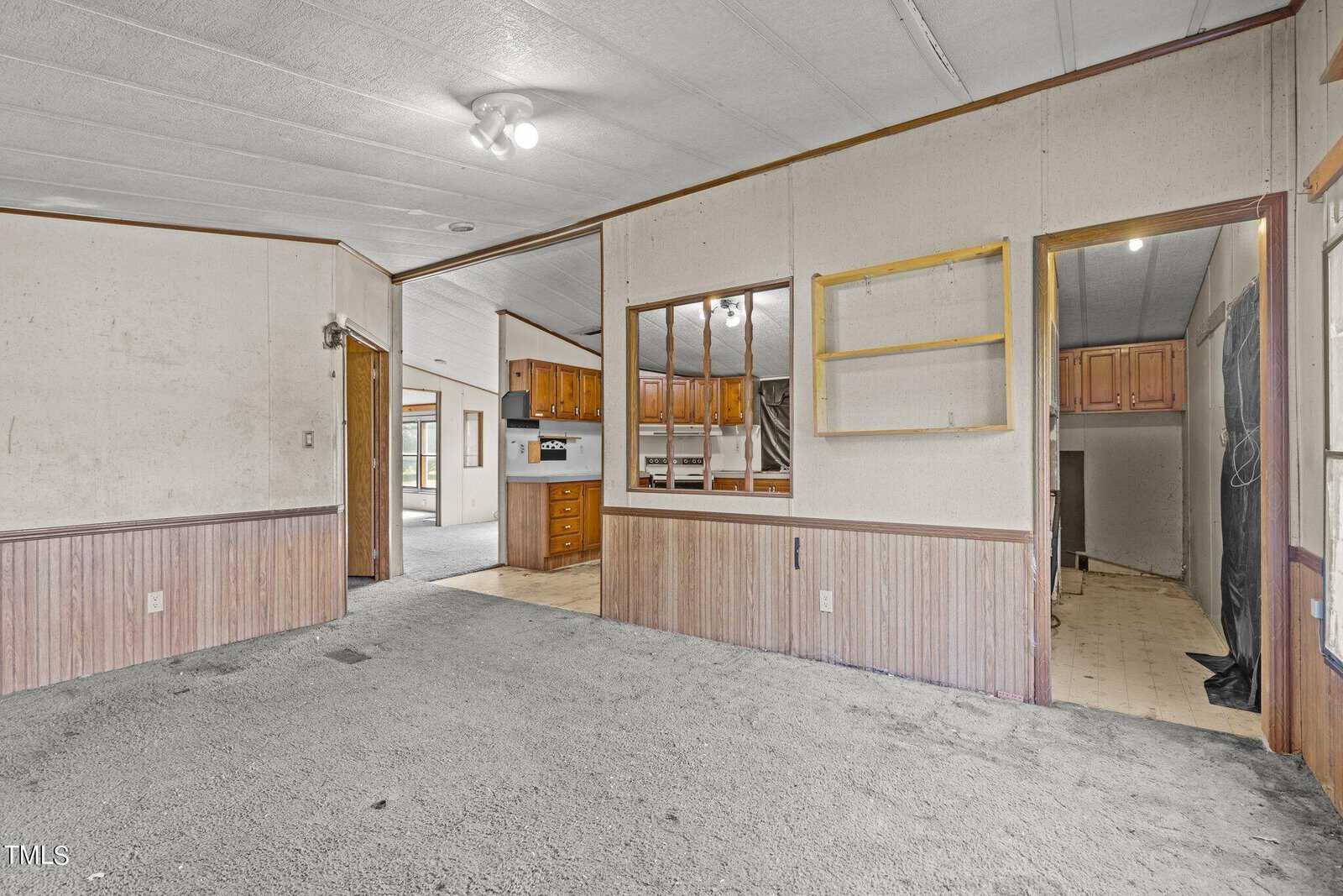 An empty room with beige walls and carpet, featuring a wooden partition with shelves. The view extends into a kitchen area with wooden cabinets and tiled flooring. Ceiling lights are visible, and a black curtain hangs to the right.