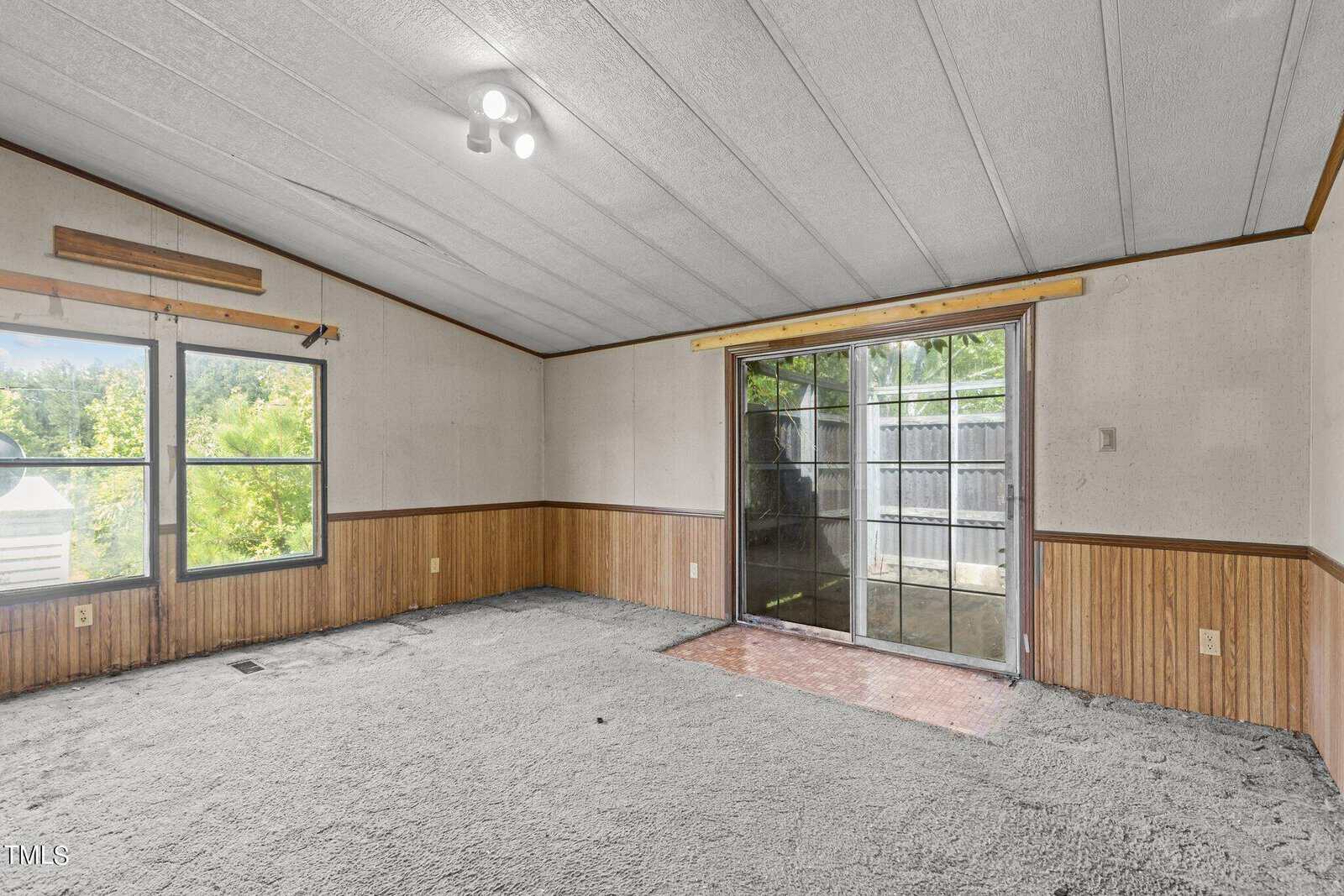 An empty room with light gray carpeting and wood paneling on the lower half of the walls. Large windows and a sliding glass door open to the outside, bringing in natural light. The ceiling has a geometric pattern and a small light fixture.