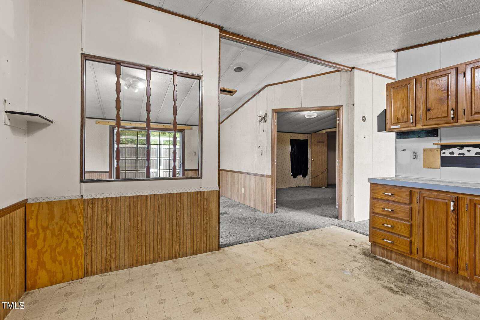 Interior of a vacant house with wood paneling and linoleum flooring. The kitchen features wooden cabinets and a white stove. An open doorway leads to a carpeted living area, with visible ceiling beams and soft lighting.