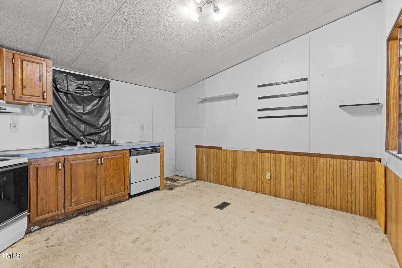A partially renovated kitchen with wooden cabinets and a white dishwasher. The floor has a worn linoleum covering, and the ceiling is light-colored. A plastic sheet is taped over a window above the sink, and there are shelving units on the walls.