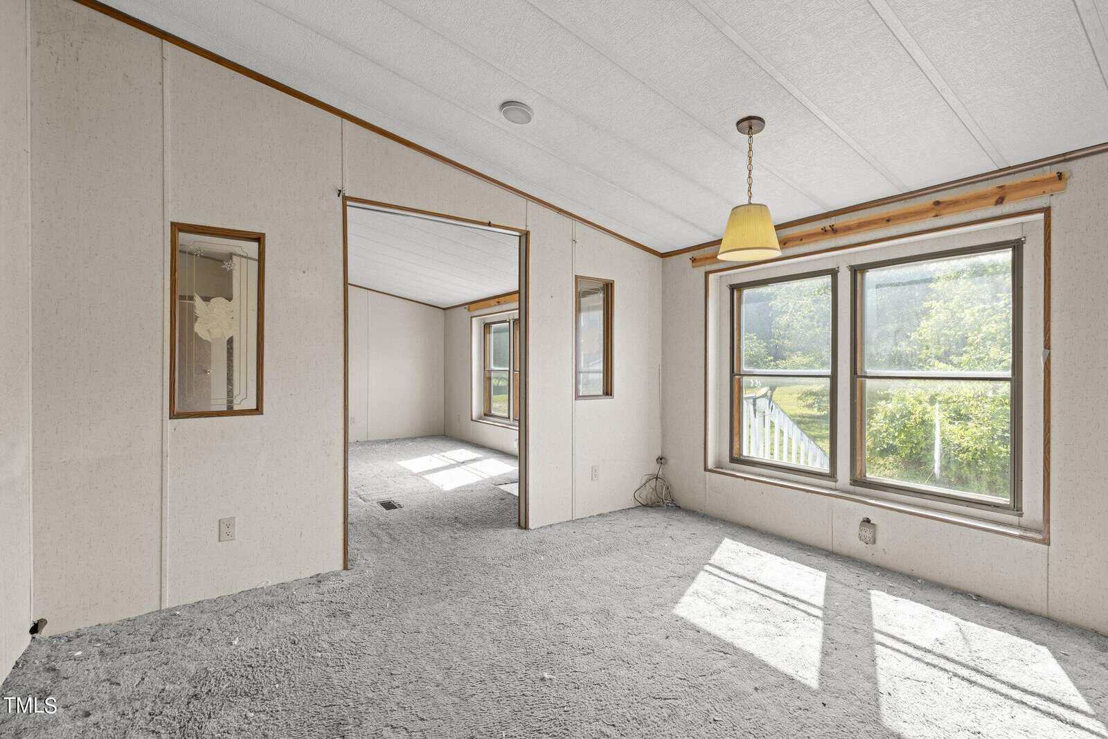 An empty room with beige walls and carpeted floor. Large windows provide natural light. A yellow hanging light fixture is in the center of the ceiling. The room opens into another space through a wide doorway.