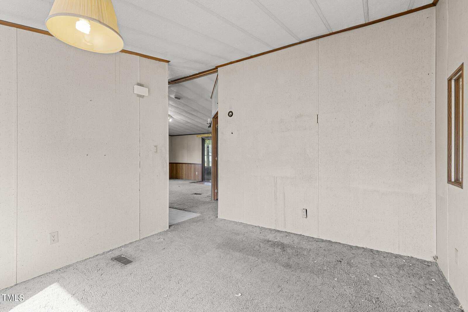 An empty room with beige walls and carpeted floor, featuring a hanging overhead light. A small window and a doorway lead to another room with wood-paneled walls. Natural light enters from the left.