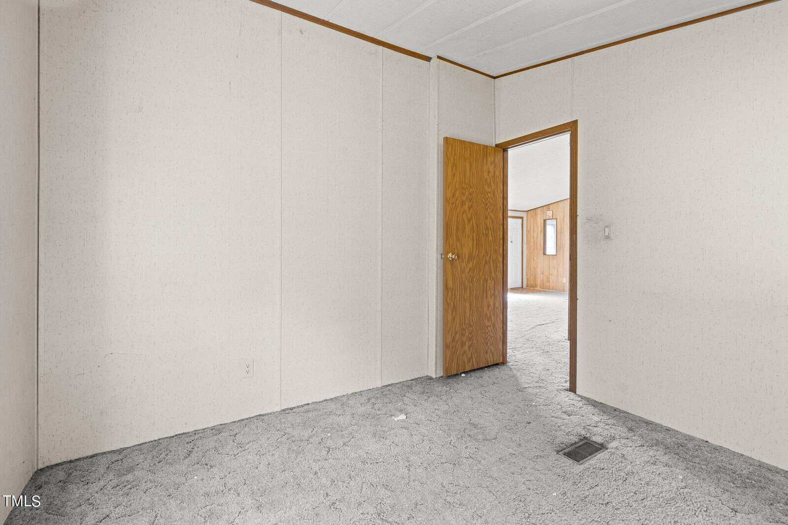 An empty room with light-colored walls and a textured carpet. The room has a wooden door that opens to another area with similar wall and carpet textures. The overall appearance is simple and undecorated.