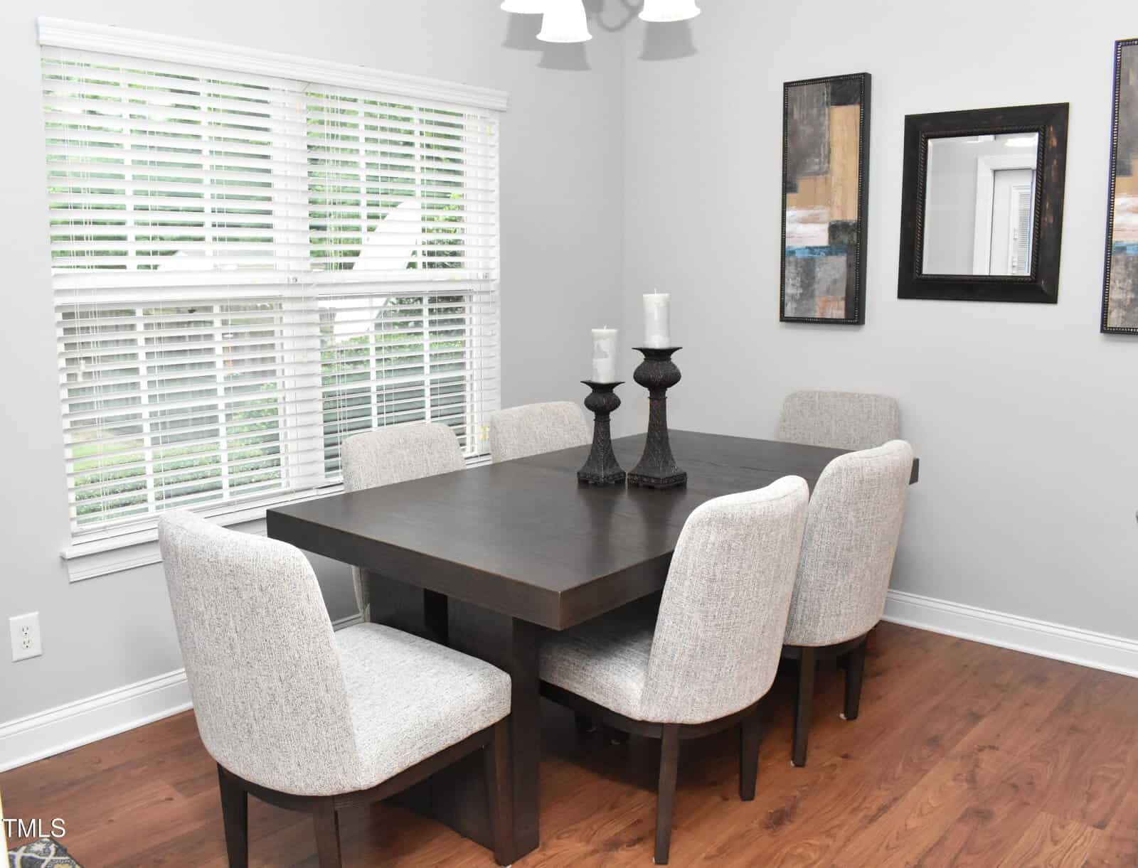 A dining room featuring a dark wooden table with six cushioned chairs. The table is topped with two decorative candle holders. The room has light walls, hardwood flooring, two abstract paintings, and a window with blinds.