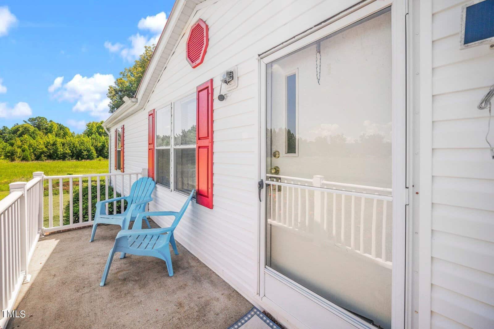 A white house with red shutters and a glass front door. Two blue Adirondack chairs sit on the porch, overlooking a grassy field and trees under a blue sky with fluffy clouds.