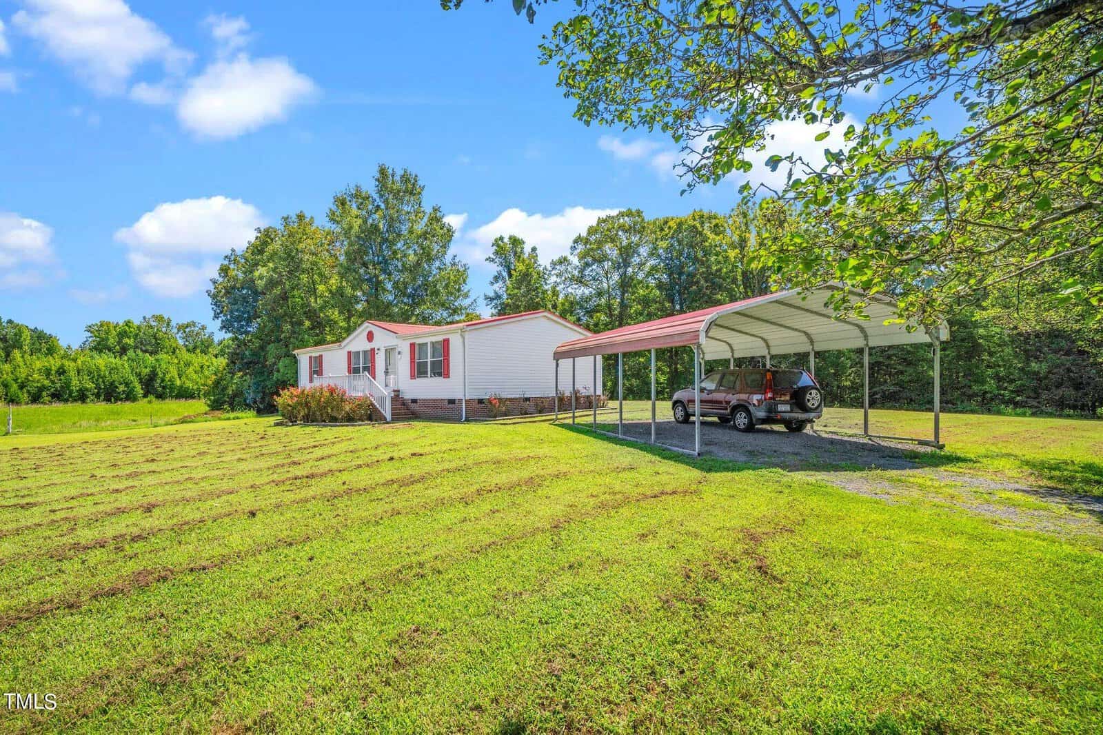 A white and red mobile home sits on a grassy lawn with a gravel driveway. A car is parked under a metal carport. Trees line the background under a bright, blue sky with scattered clouds.