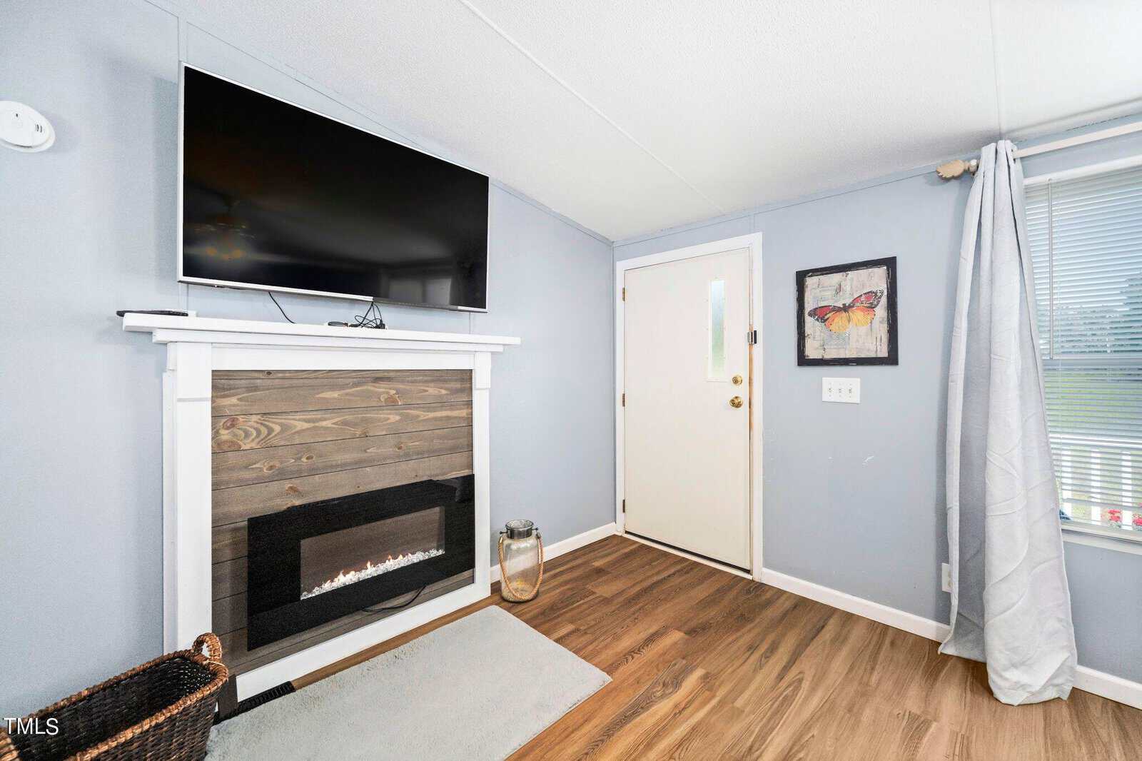 Room with a light blue wall and wood flooring, featuring a wall-mounted TV above a wooden fireplace. A door with a window is next to a framed butterfly picture. A gray curtain covers a window on the right, and a woven basket sits on a rug.