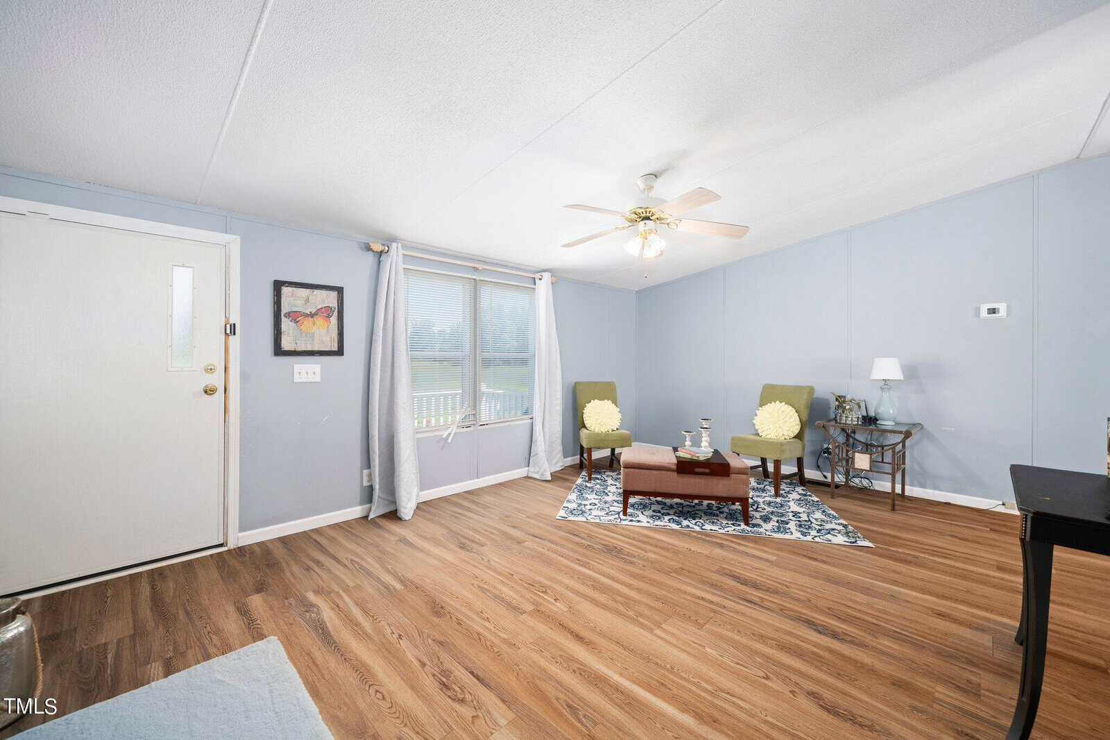 A spacious living room with light blue walls and wooden flooring. It features a ceiling fan, a large window with curtains, a small table with a lamp, two green chairs, a patterned rug, and decorative wall art.