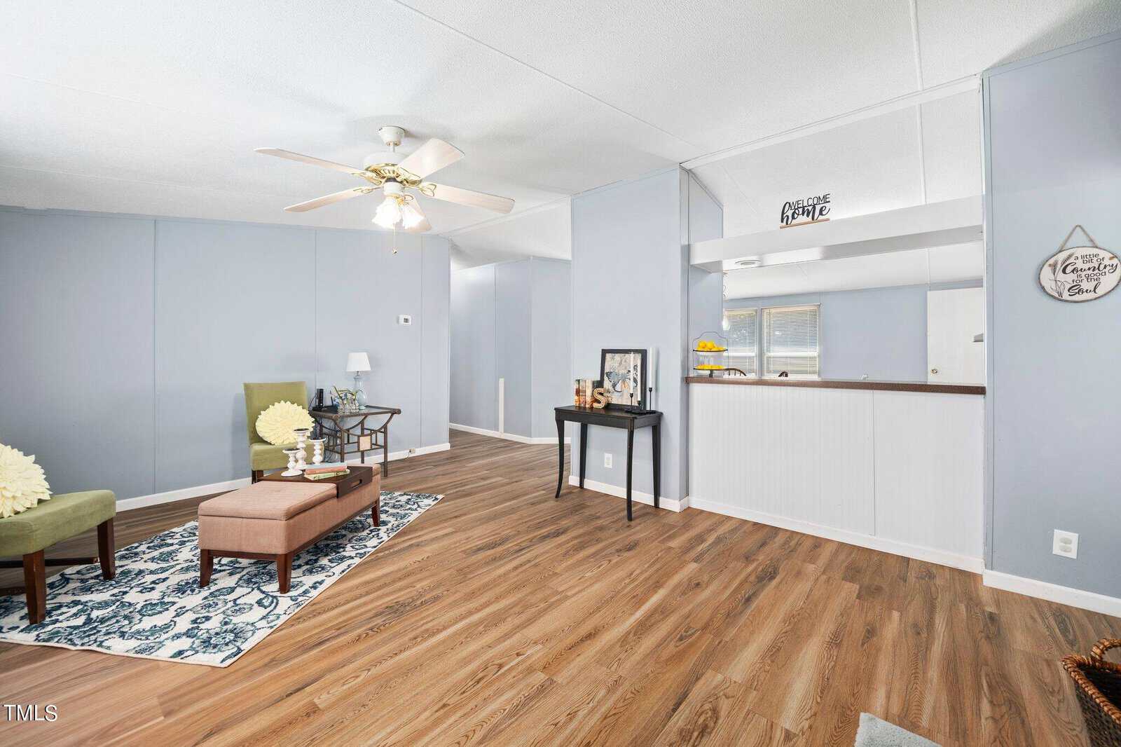 A cozy living room with light blue walls and wooden floor. It features a brown ottoman, green chair, a small table with decor, and a ceiling fan. A narrow hallway leads to other rooms, with a window visible in the background.