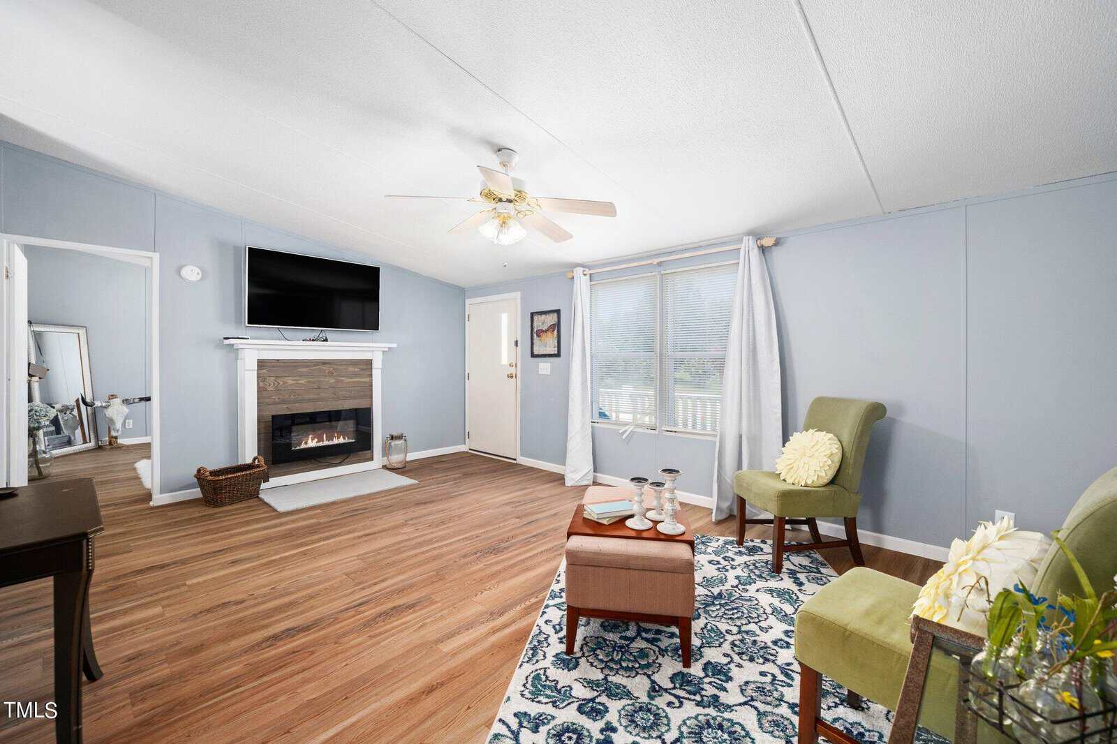 Bright living room with light blue walls and wood flooring. Features a fireplace with a TV above, two green chairs with a floral rug, and a small table. A ceiling fan hangs above, and a large window lets in natural light.