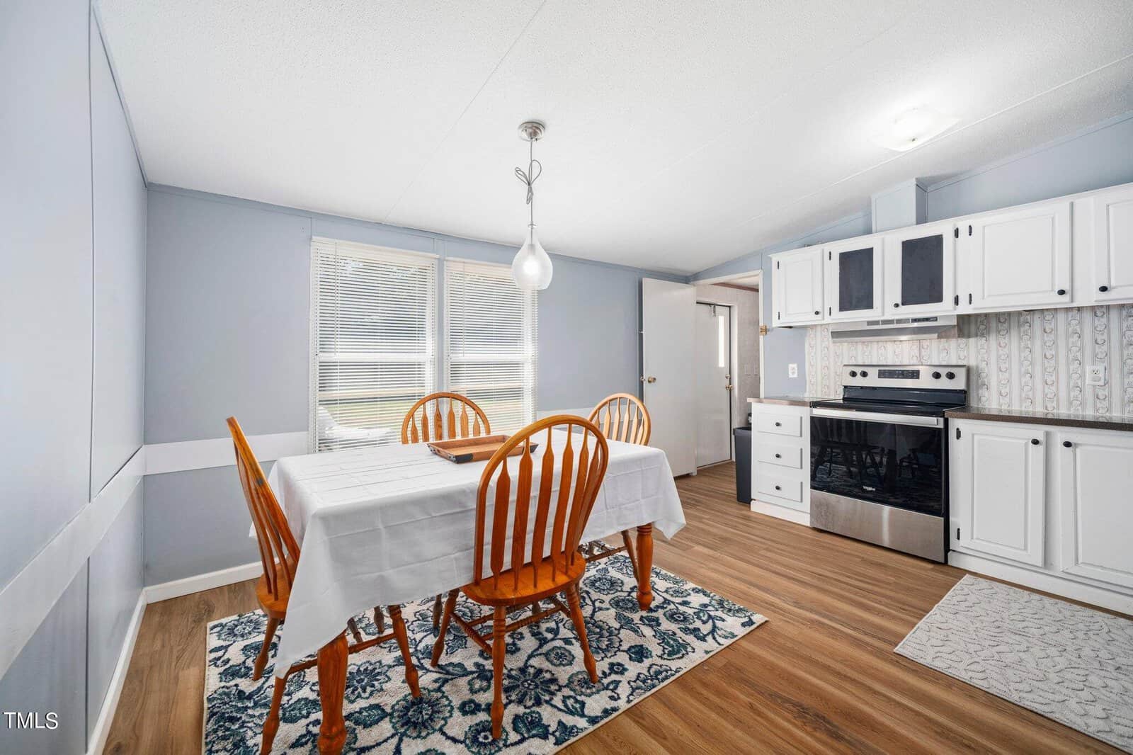A kitchen with light blue walls, featuring a wooden dining table covered with a white tablecloth and four matching chairs. The room has a stove, white cabinets, and a patterned rug on a wood floor. A pendant light hangs above the table.
