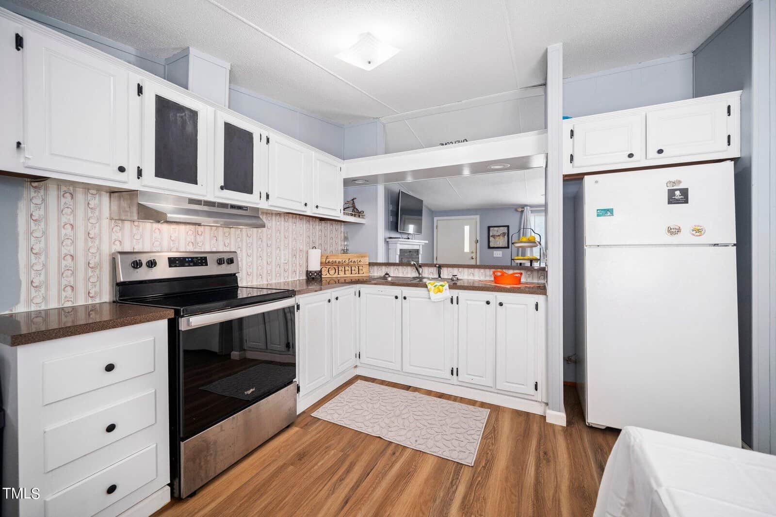 A modern kitchen with white cabinets, a stainless steel oven, and a white refrigerator. Wooden flooring enhances the space. A countertop holds decorative items, including a basket and an orange pot. A patterned mat lies in front of the oven.