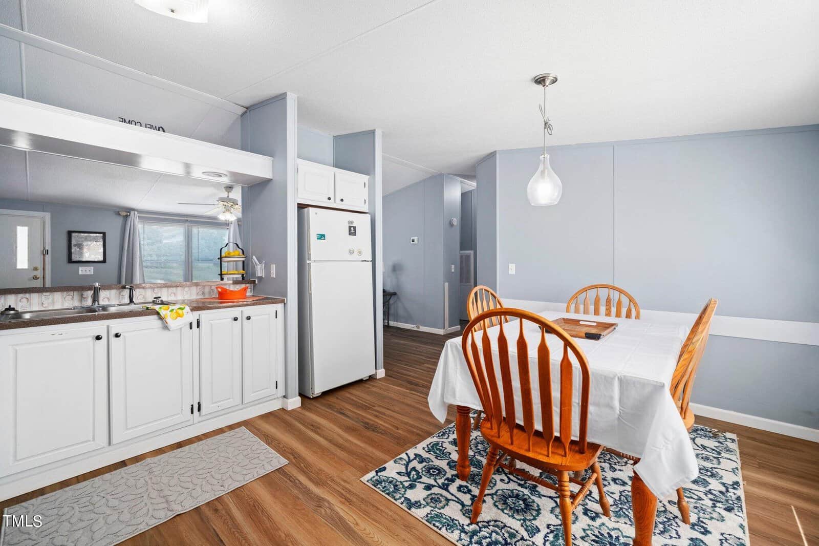 A modern dining area with a wooden table and six chairs set on a patterned rug. A white cabinet and refrigerator are visible, along with pendant lighting. The room has light blue walls and wooden flooring. Natural light enters through a window.