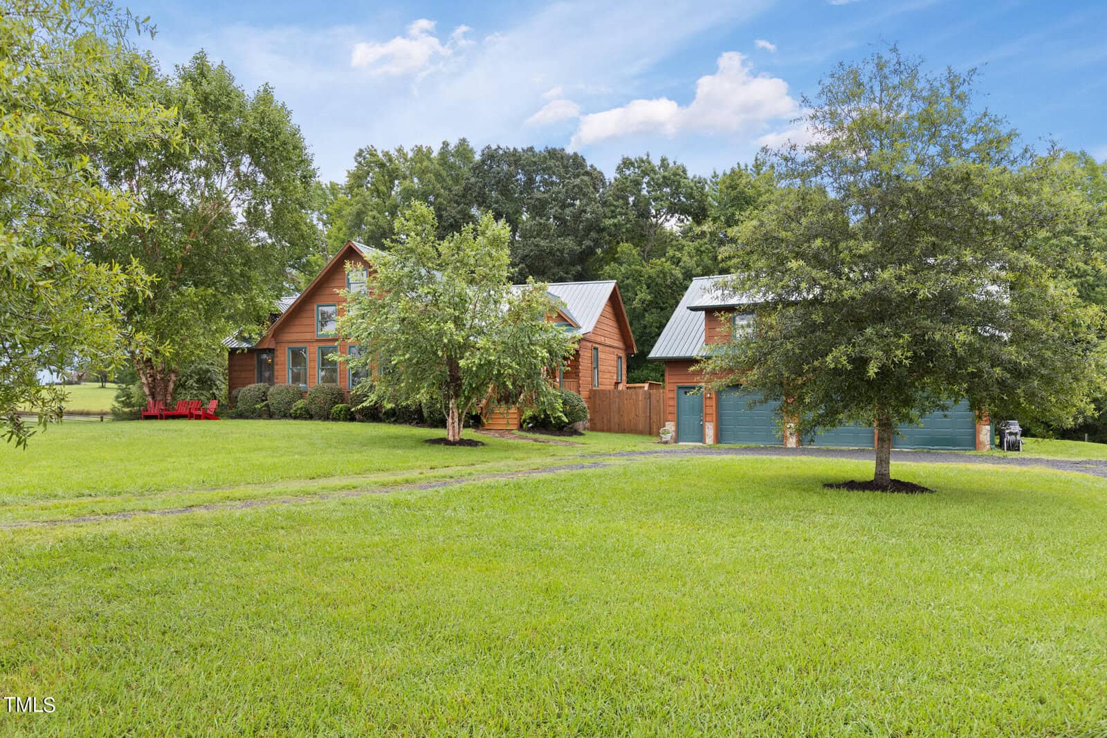 A rustic wooden house with a metal roof sits amidst a lush green lawn and trees under a blue sky. A matching detached garage is nearby, with a gravel driveway leading to it. Bushes and shrubs surround the house, adding to the serene countryside setting.
