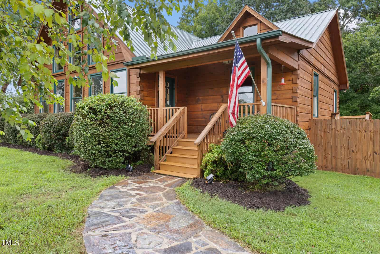 A charming wooden cabin with a small porch sits surrounded by lush greenery. An American flag is displayed on the porch. A stone pathway leads to the front door, flanked by neatly trimmed bushes.
