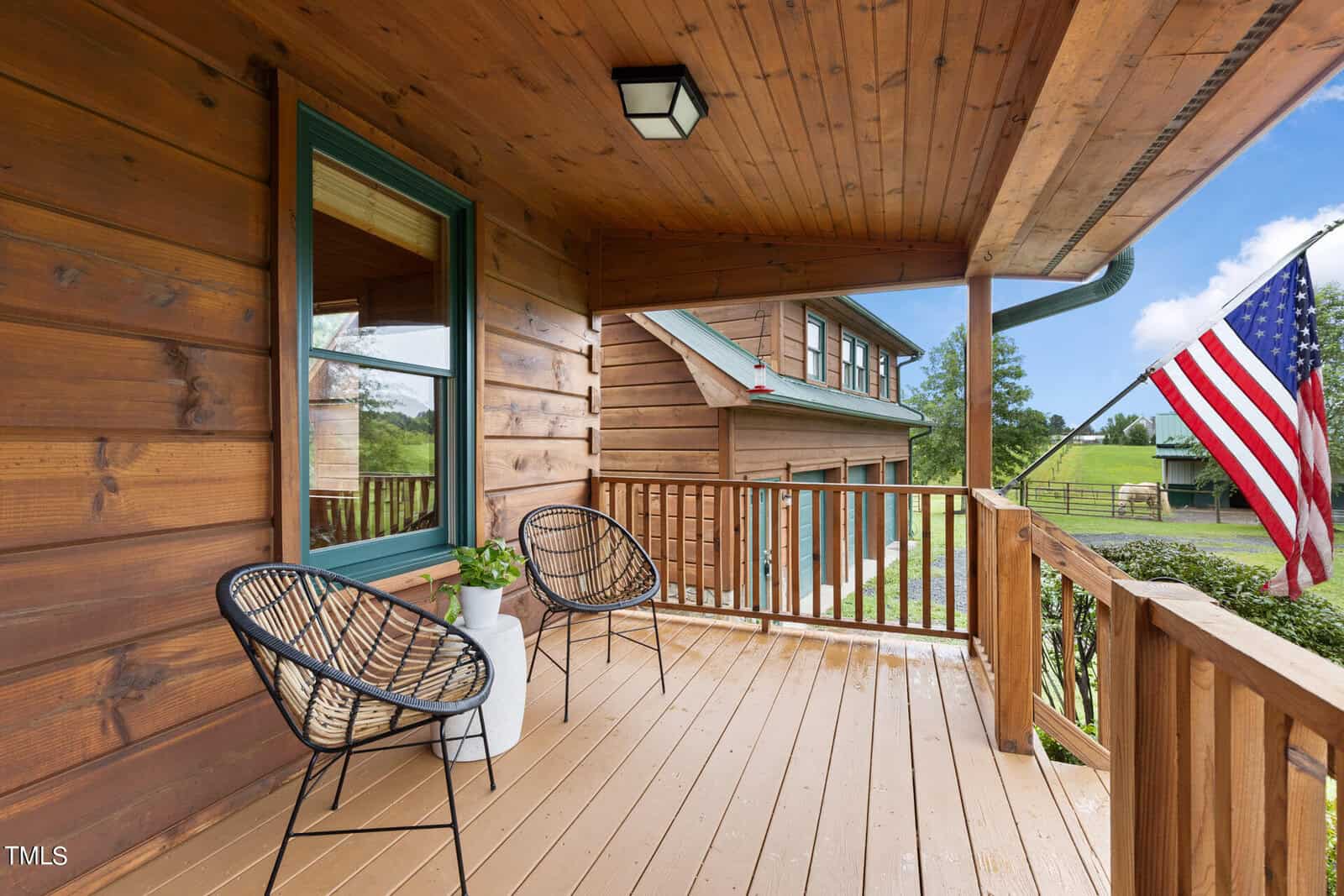 A wooden porch featuring two modern black wire chairs and a white planter. An American flag is displayed on the railing. The porch overlooks a grassy landscape with buildings in the background under a partly cloudy sky.