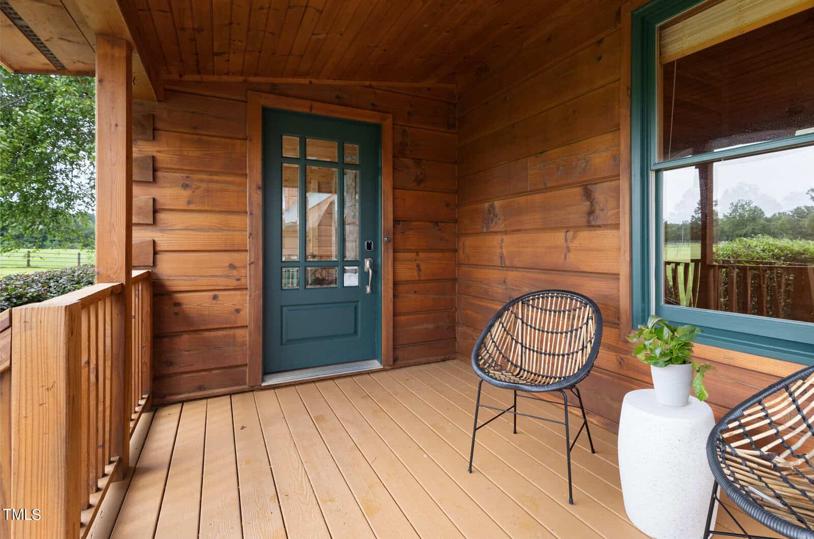 A cozy wooden porch featuring a green door, two black wicker chairs, and a small white table with a potted plant. The porch overlooks a green landscape with trees, under a wooden ceiling.