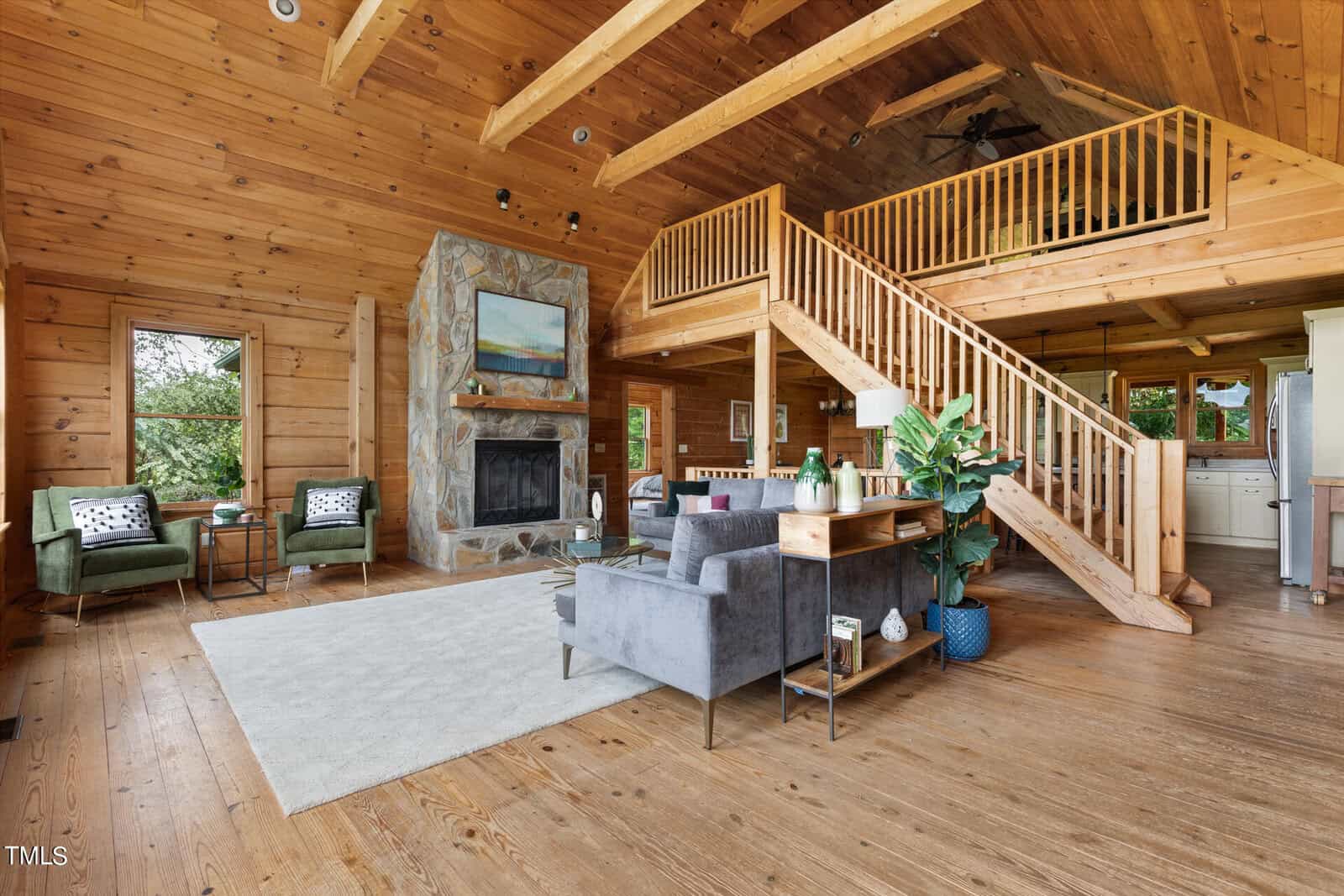 Spacious wooden cabin interior with vaulted ceiling, featuring a stone fireplace, gray sofa, green armchairs, and a rug. A wooden staircase leads to an upper loft, and large windows reveal green foliage outside.