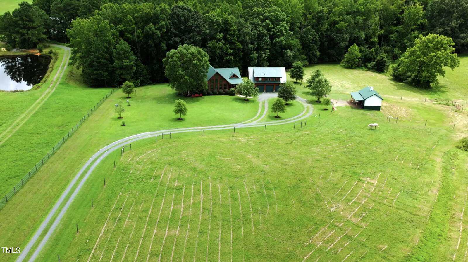 Aerial view of a rural landscape with a winding driveway leading to a rustic house surrounded by lush green fields and trees. A small barn sits nearby, and a fenced area contains a few animals. A pond is visible on the left.