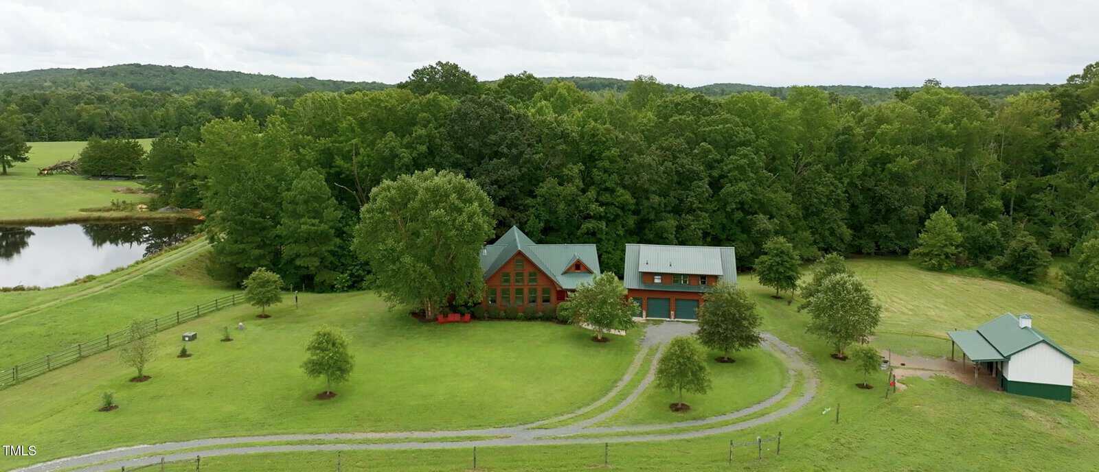 Aerial view of a large, green property featuring a rustic house with a green roof surrounded by lush trees. A curved driveway leads to the house. A small pond is visible to the left and a shed-like structure is on the right.