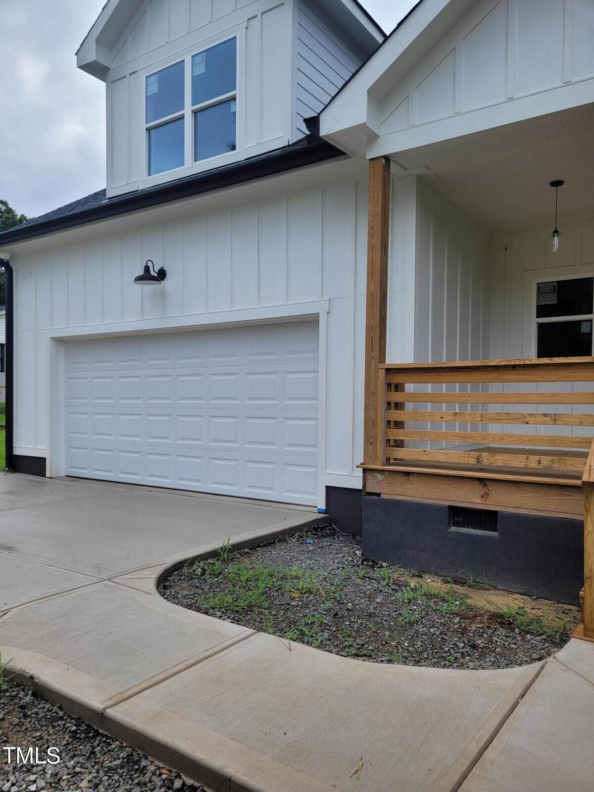 Modern two-story house with white siding and a black roof. Features a two-car garage and a wooden porch with railing. The driveway and walkway are paved, with a small patch of greenery near the entrance.