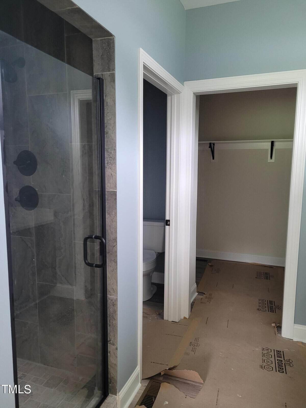 A bathroom with a modern glass shower on the left and a toilet in a separate small room straight ahead. The floor is covered with protective cardboard, suggesting ongoing construction or renovation.