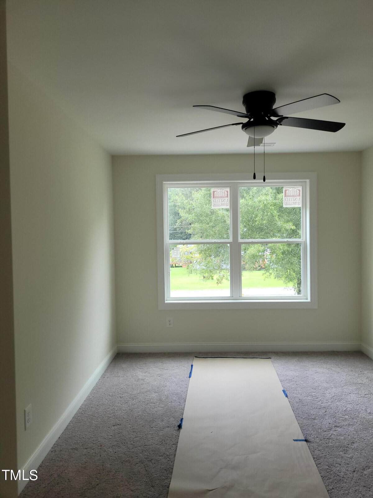 A bright, unfurnished room with light-colored walls and carpeted flooring. A large window with tree views lets in natural light. A ceiling fan is centered above, and a sheet of paper is laid out on the floor.