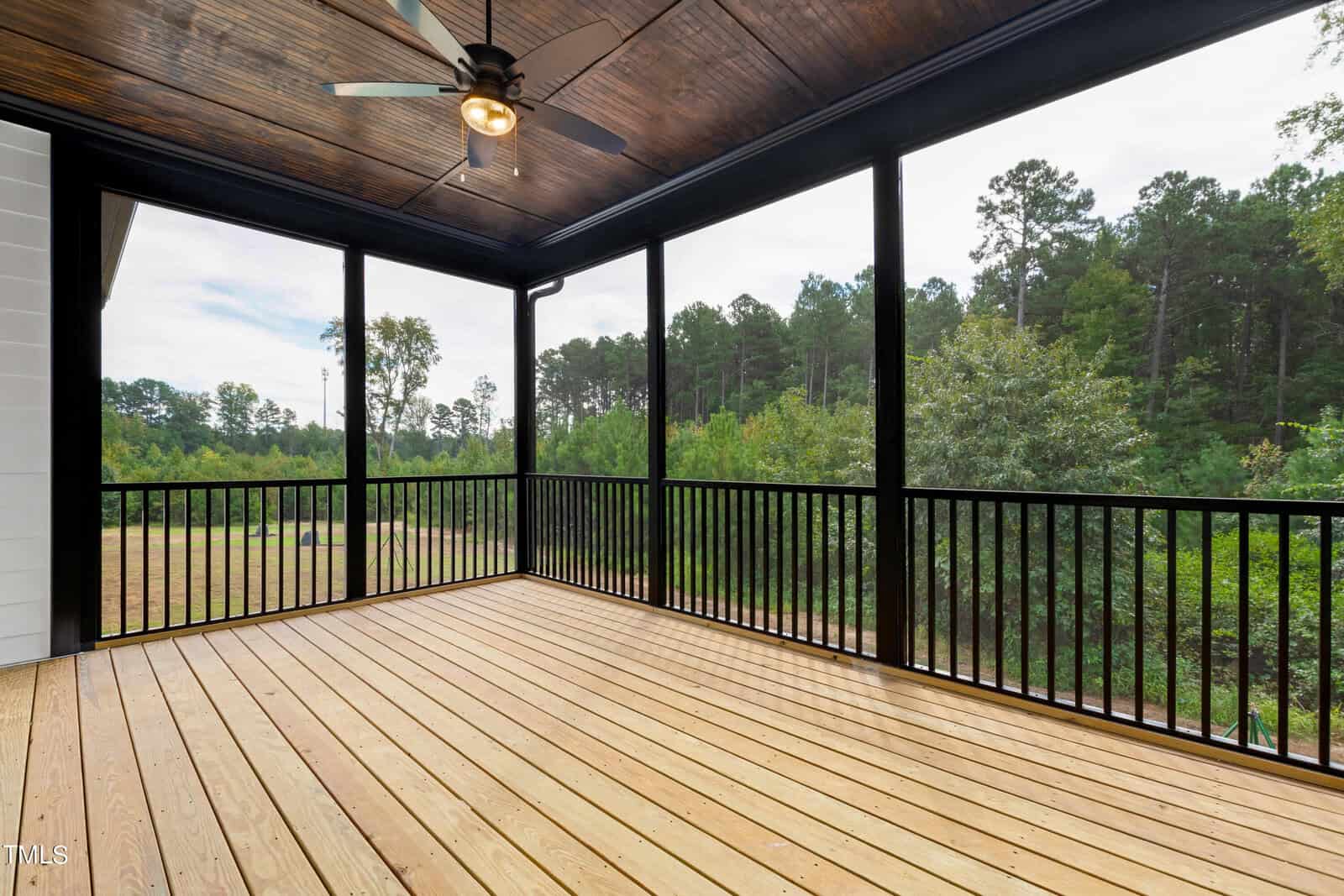 Screened-in porch with wooden flooring and a dark wooden ceiling featuring a ceiling fan. The porch overlooks a grassy yard bordered by a dense area of trees. Black railings surround the space.
