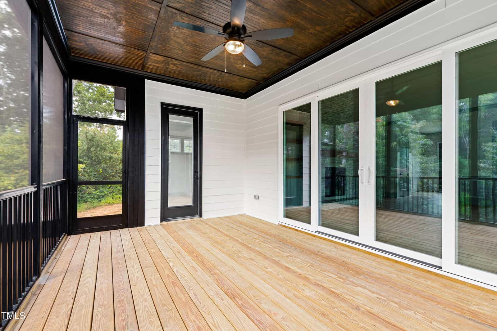 A covered wooden deck with a ceiling fan and recessed lighting. The white-paneled walls contrast with the dark ceiling. Large sliding glass doors and a single glass door open to the outside, with greenery visible through screened sections.