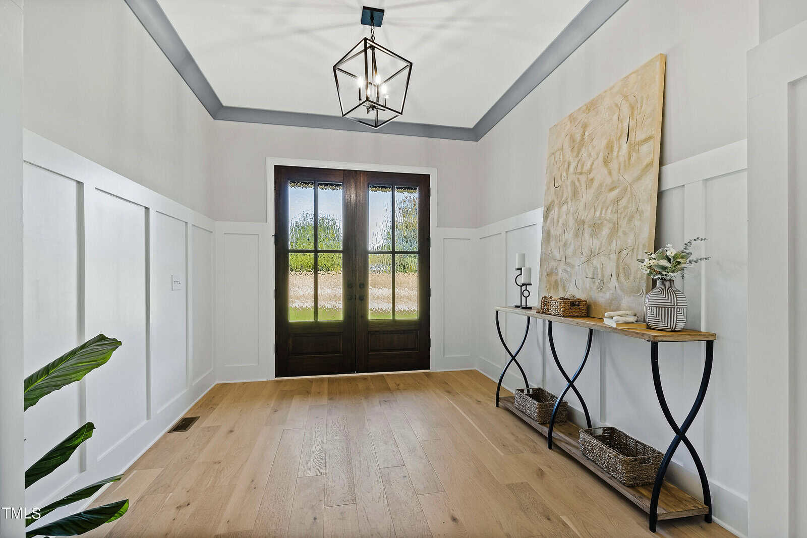 A hallway with light wood flooring and white paneled walls features a dark wooden front door with glass panes. A narrow console table holds decorative items, including a large textured artwork, wicker baskets, a candle, and a vase with flowers.