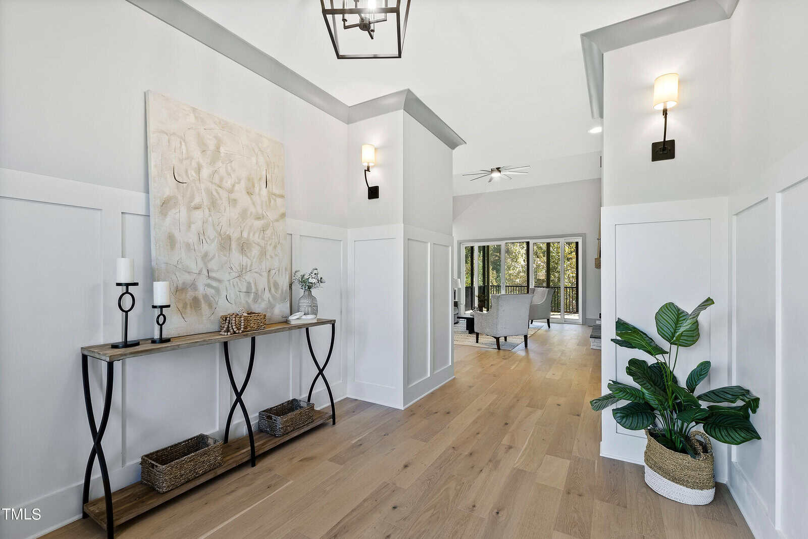 A modern hallway with white walls and light wood flooring. A wooden console table holds candles and decor. Potted plants are on the floor. Wall sconces provide lighting. The end of the hallway opens to a seating area with large windows.