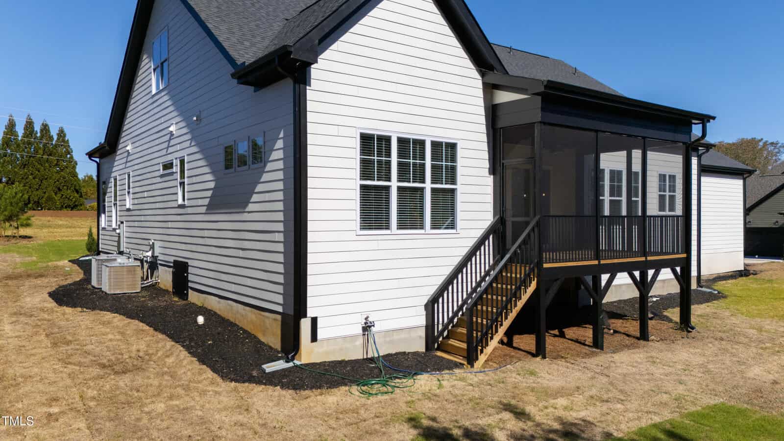 A two-story house with white siding and a dark roof is shown. It has multiple windows and an elevated screened porch with stairs leading down to the grass. The surrounding yard appears dry with patches of grass.