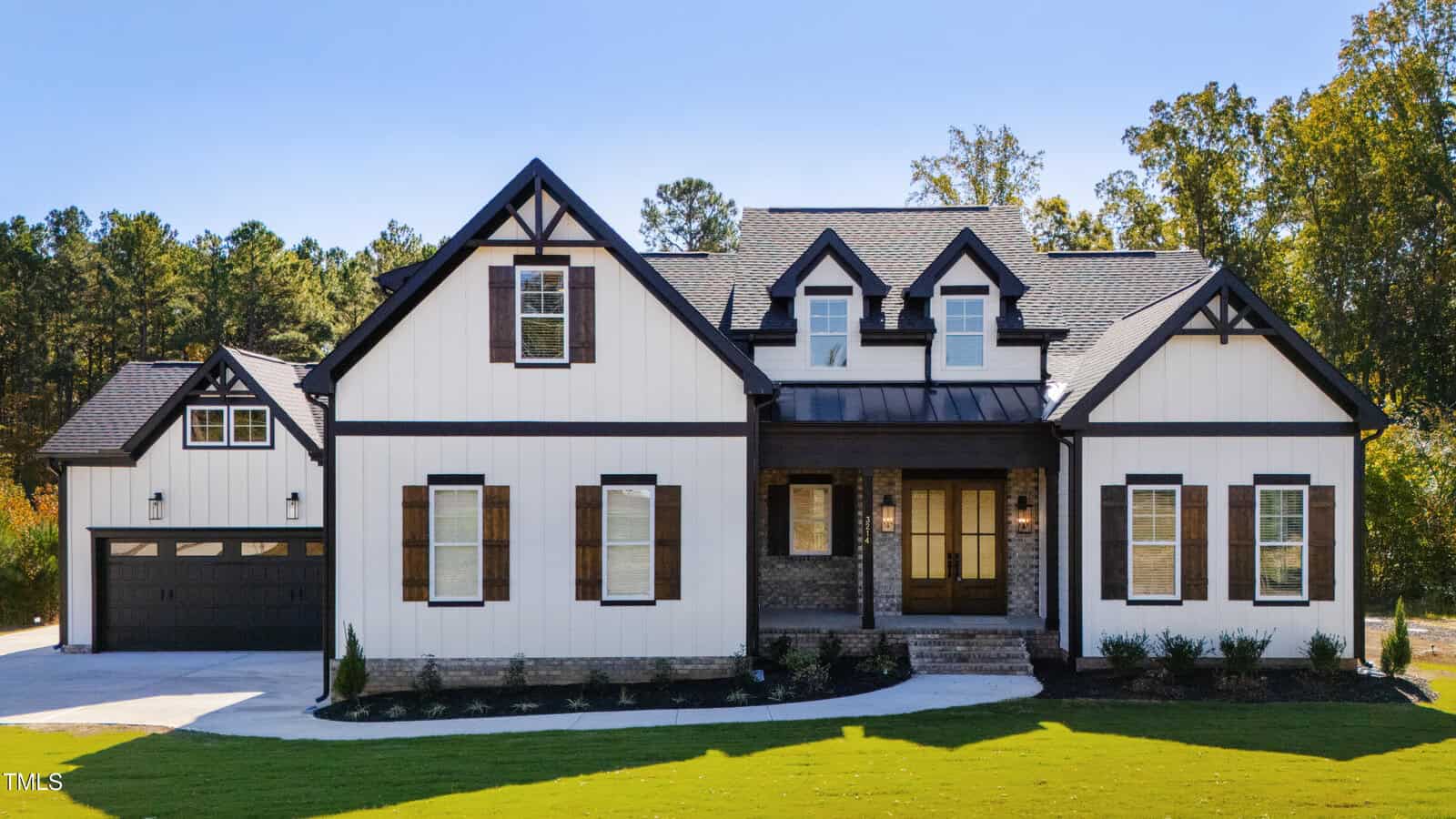 A large two-story house at 3214 Bruce Garner Rd boasts a black roof, white siding, and dark trim. It features multiple gables, a two-car garage, and a well-maintained front lawn in scenic Creedmoor. Trees grace the background under a clear blue sky.