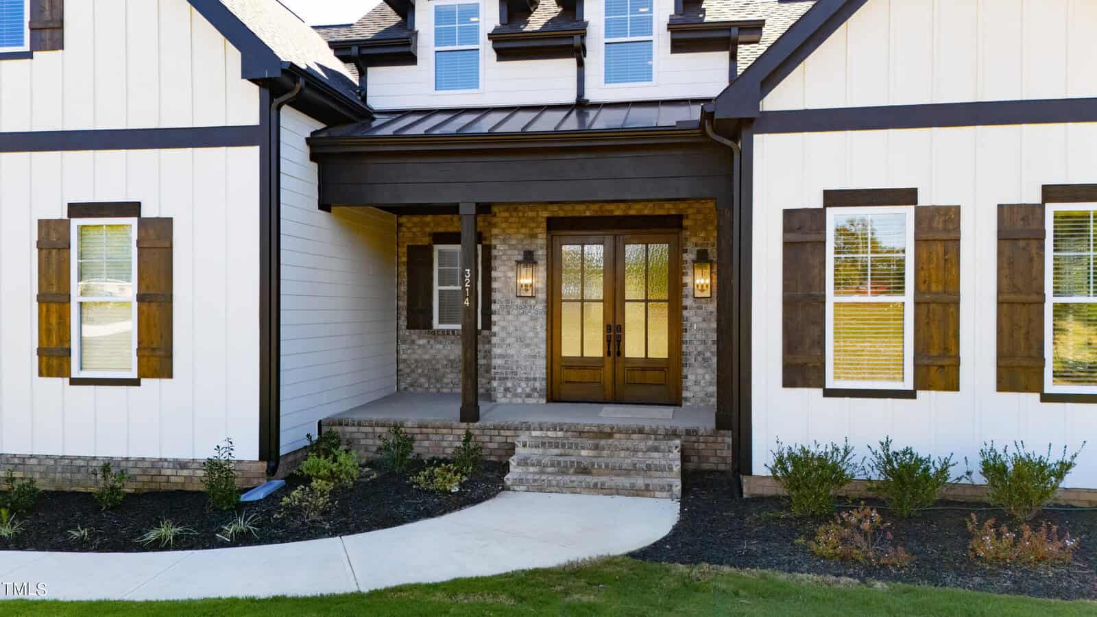 Front view of a house with a dark wooden double door entrance. The house features white siding with dark brown wooden accents and shutters. There are three small front steps leading to a covered porch with two wall lanterns. Shrubs line the walkway.