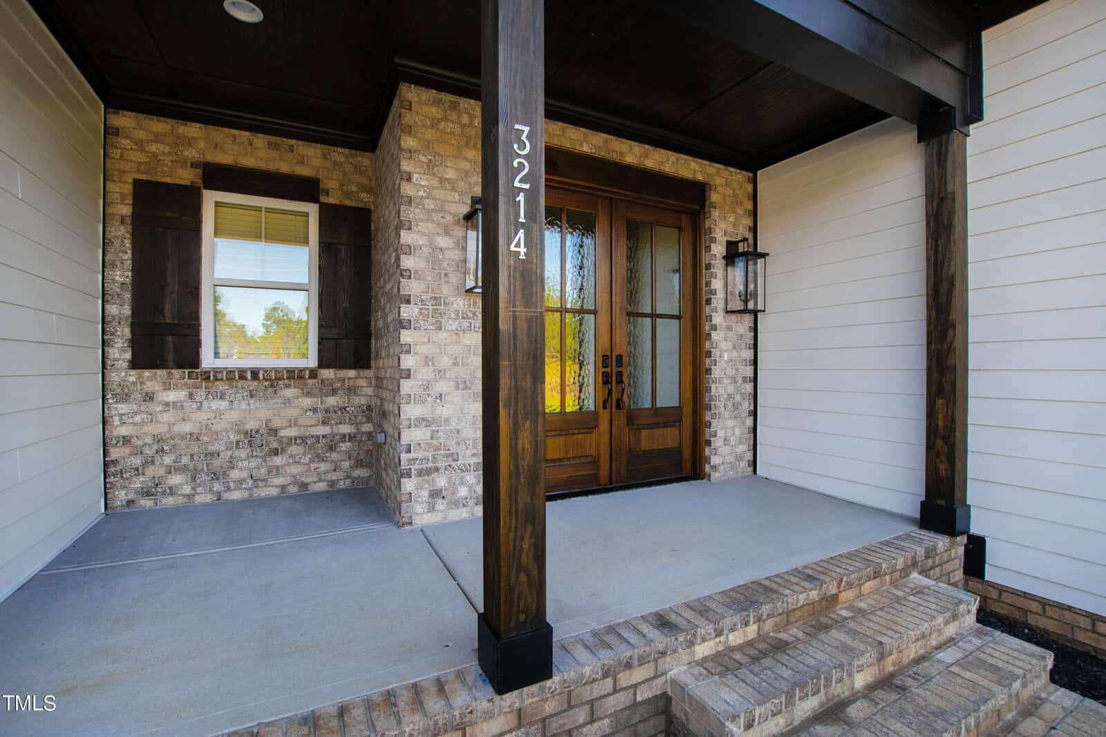 The image shows a brick and wood front porch with a large wooden door with glass panels. The house number 3214 is displayed on a wooden column. There is one window with shutters to the left of the door and two wall-mounted lantern-style lights.