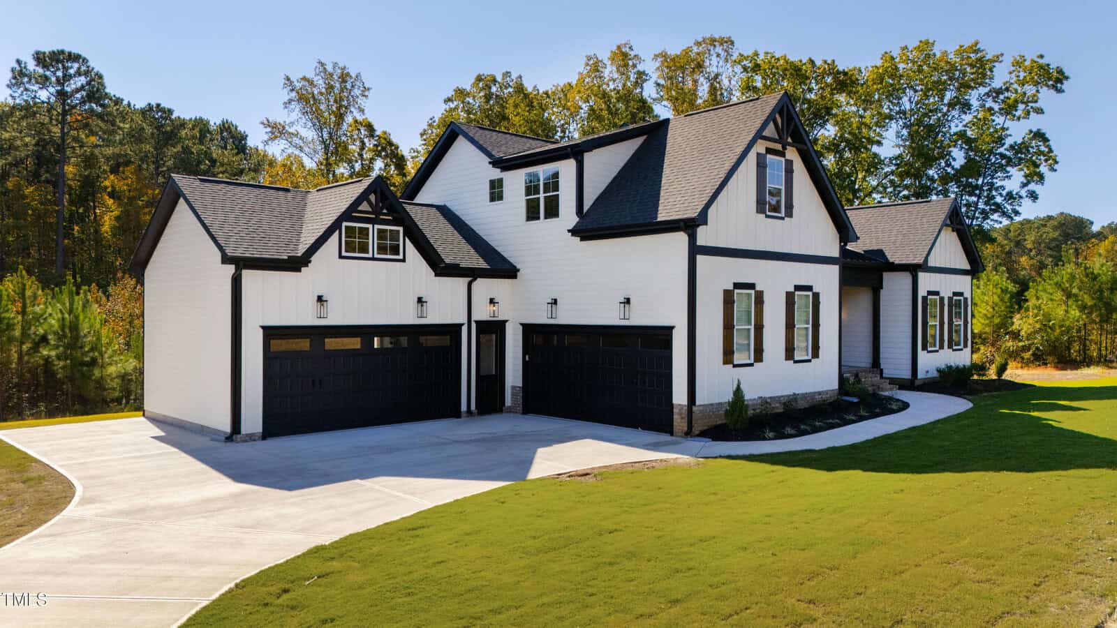 A modern two-story white house with black trim features a triple garage and a paved driveway. It is surrounded by a neatly landscaped lawn and bordered by trees. The sky is clear, indicating a sunny day.
